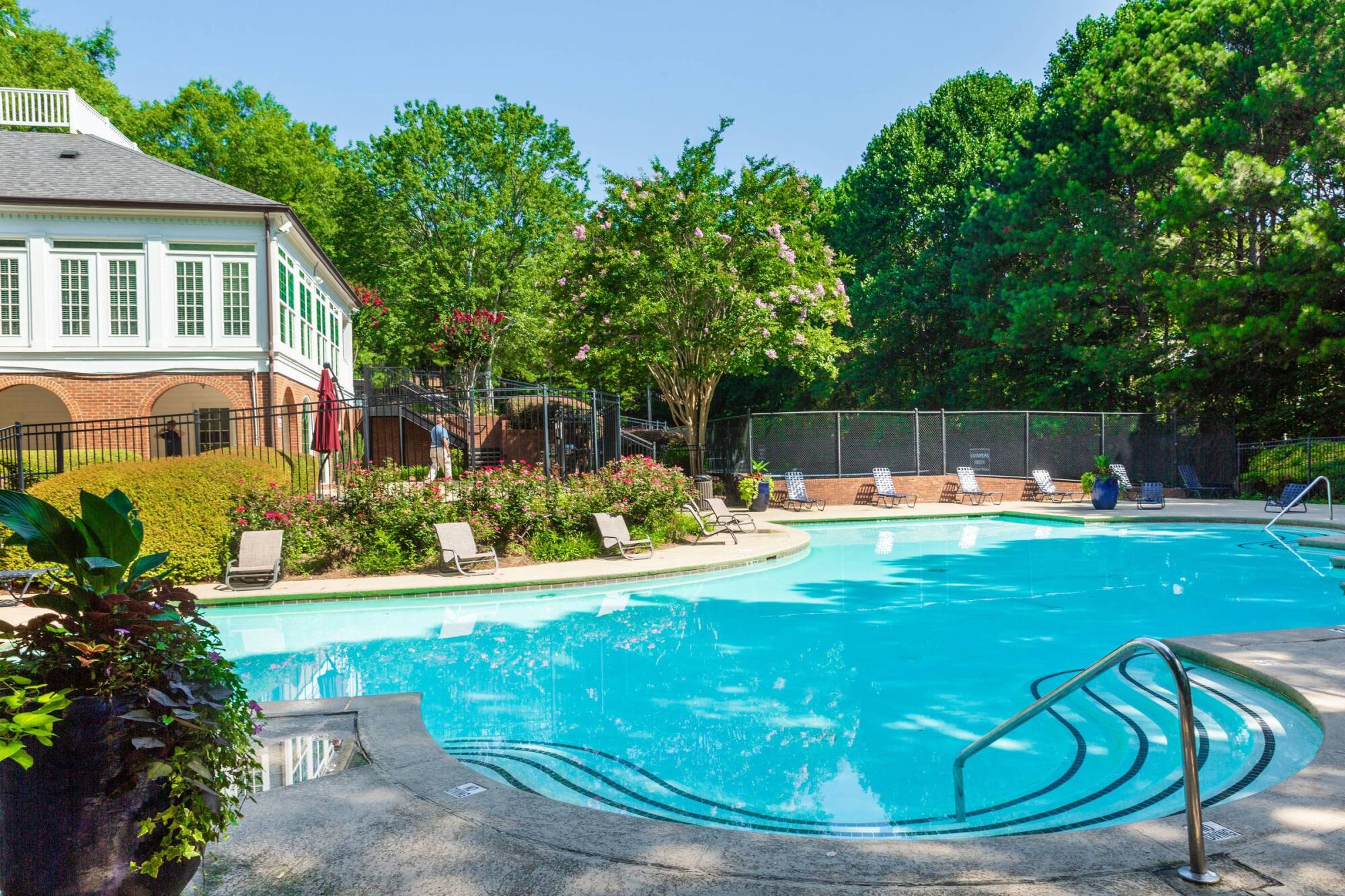 Outdoor pool with lounge chairs in front of a brick and white building, surrounded by trees on a sunny day.