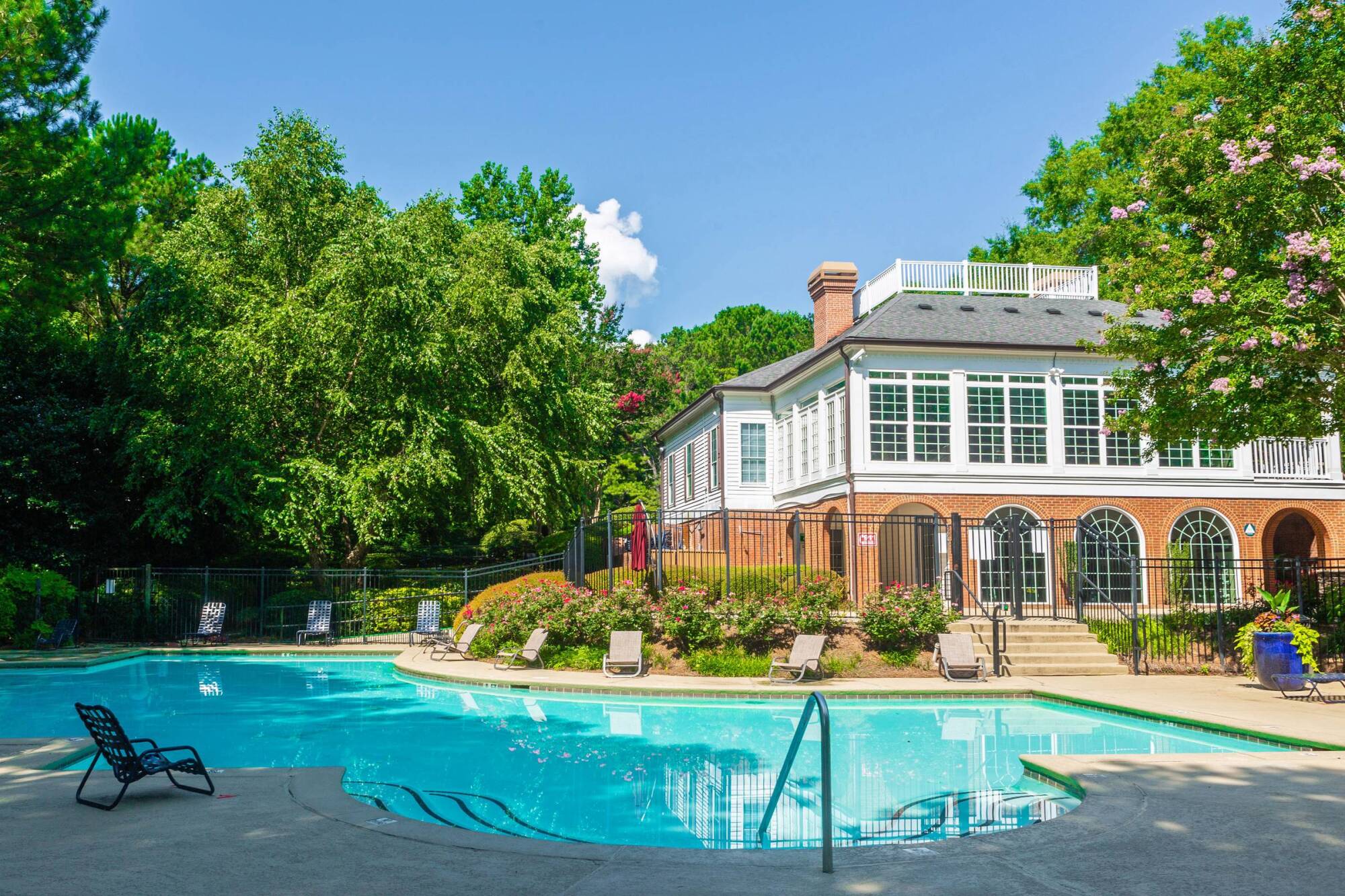 Outdoor pool with lounge chairs in front of a brick and white building, surrounded by trees on a sunny day.