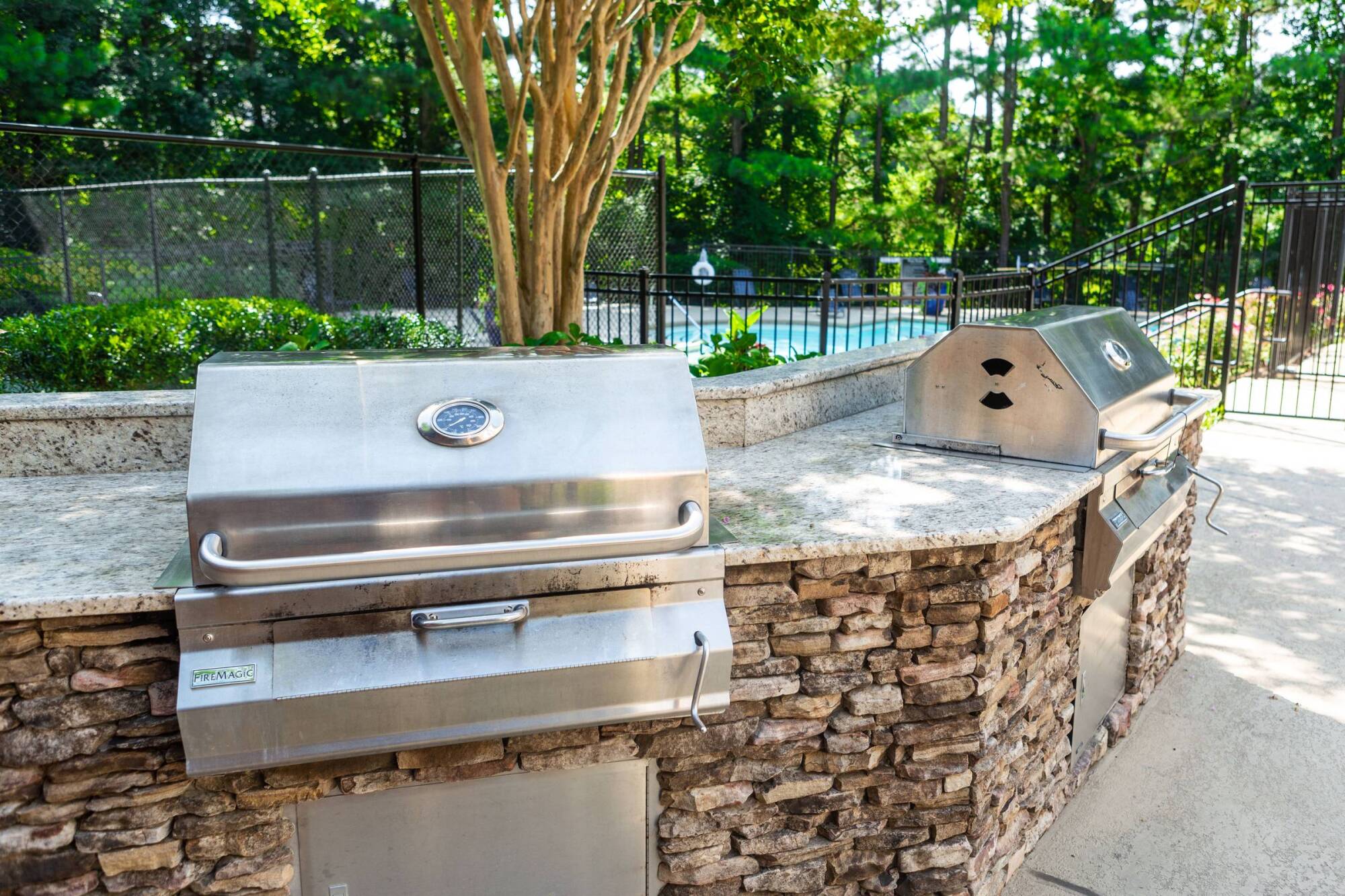 Stainless steel outdoor grill on a stone counter near a pool, surrounded by trees and greenery.