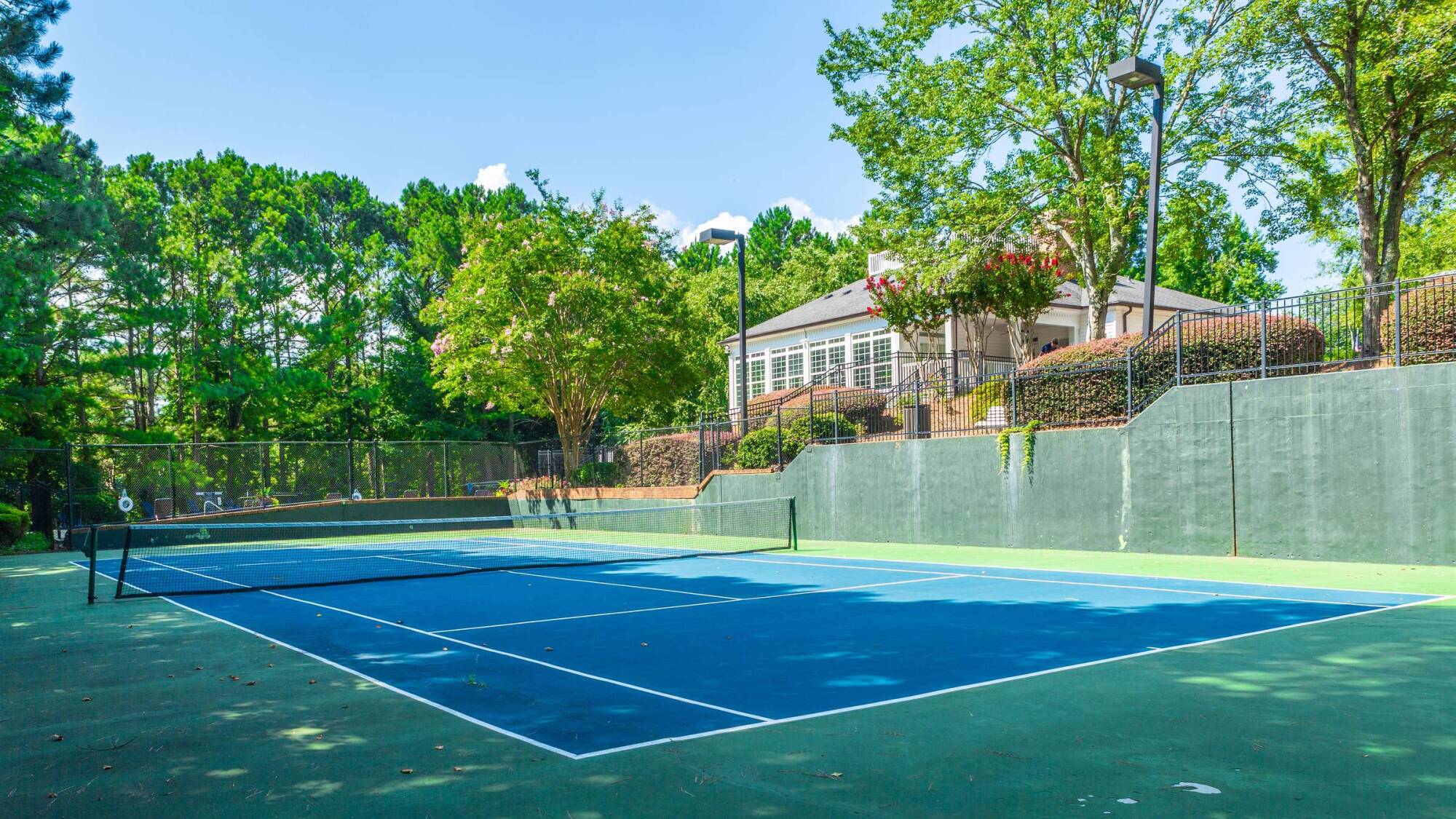 Outdoor tennis court with a blue surface, surrounded by trees and a building in the background on a sunny day.