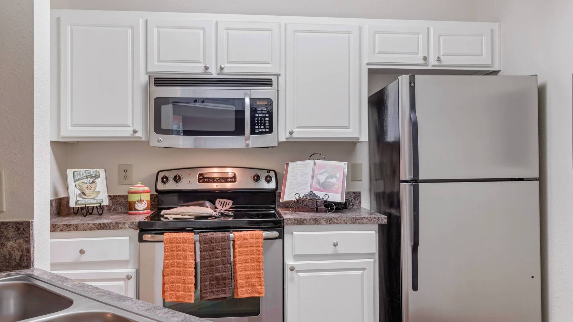 A kitchen with white cabinets, a stove, a microwave, a fridge, and orange and brown towels on the oven.