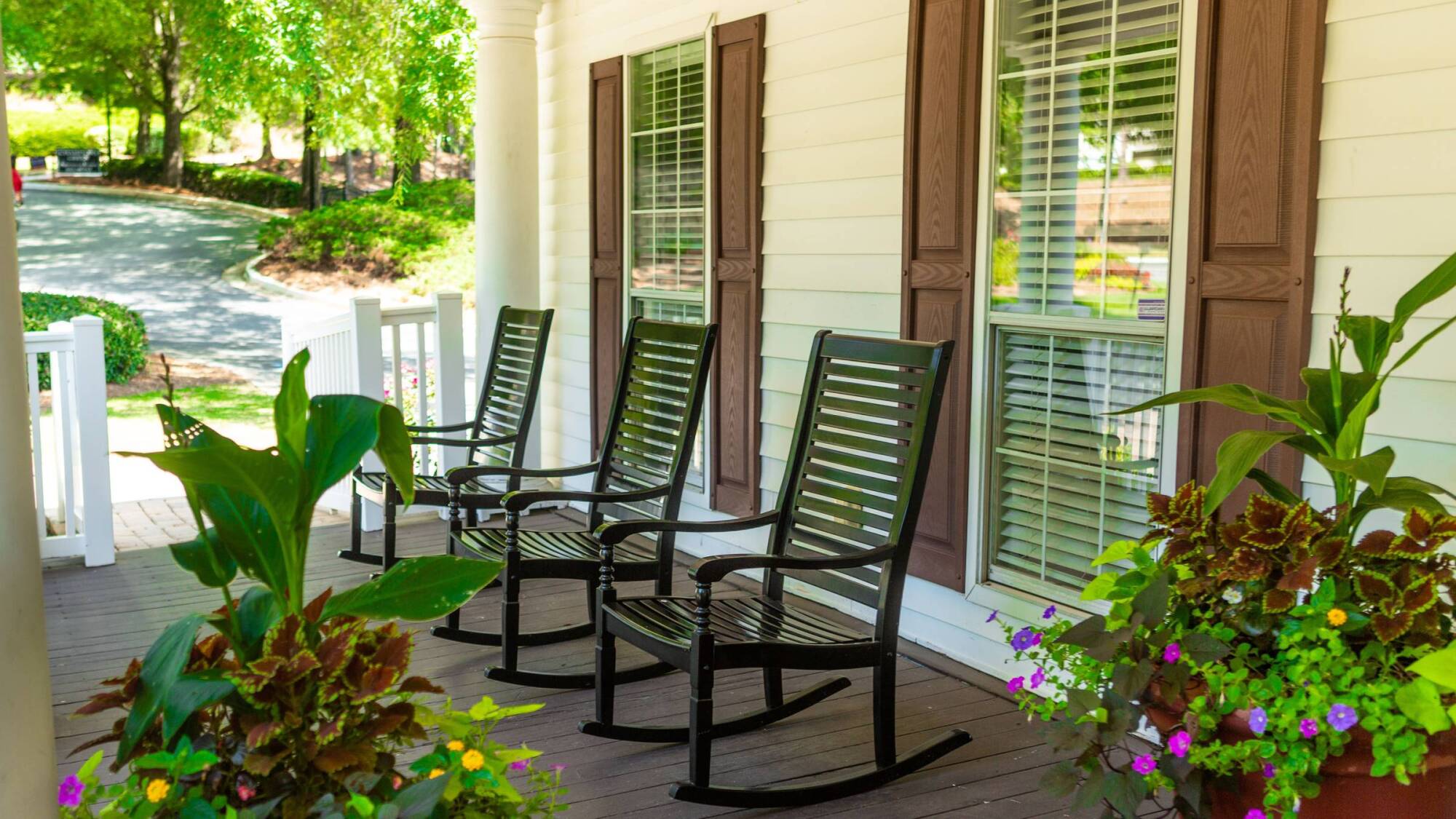 Three black rocking chairs sit on a front porch with brown shutters and potted plants nearby.