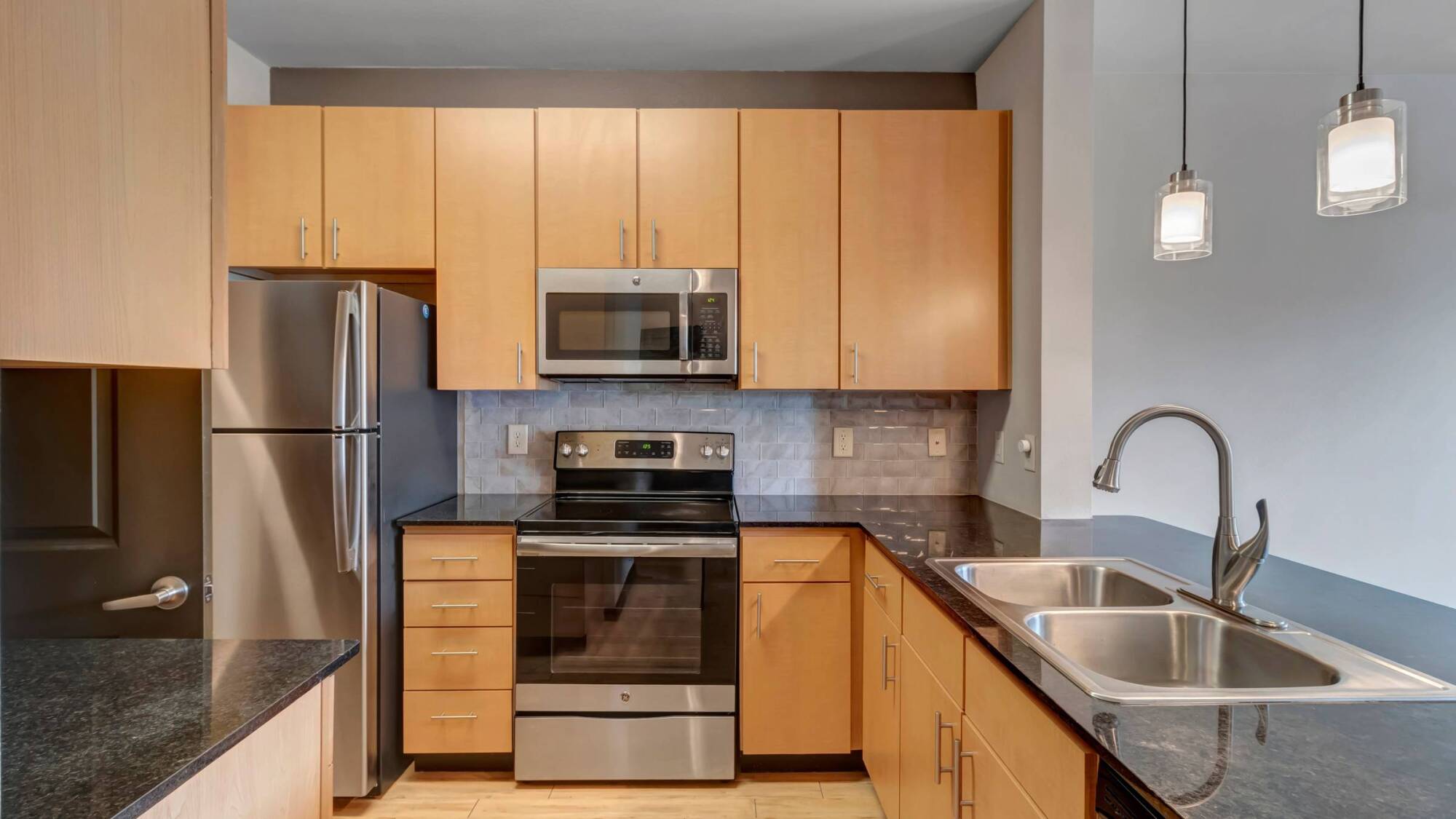 Modern kitchen with light wood cabinets, stainless steel appliances, and a double sink with a black countertop.