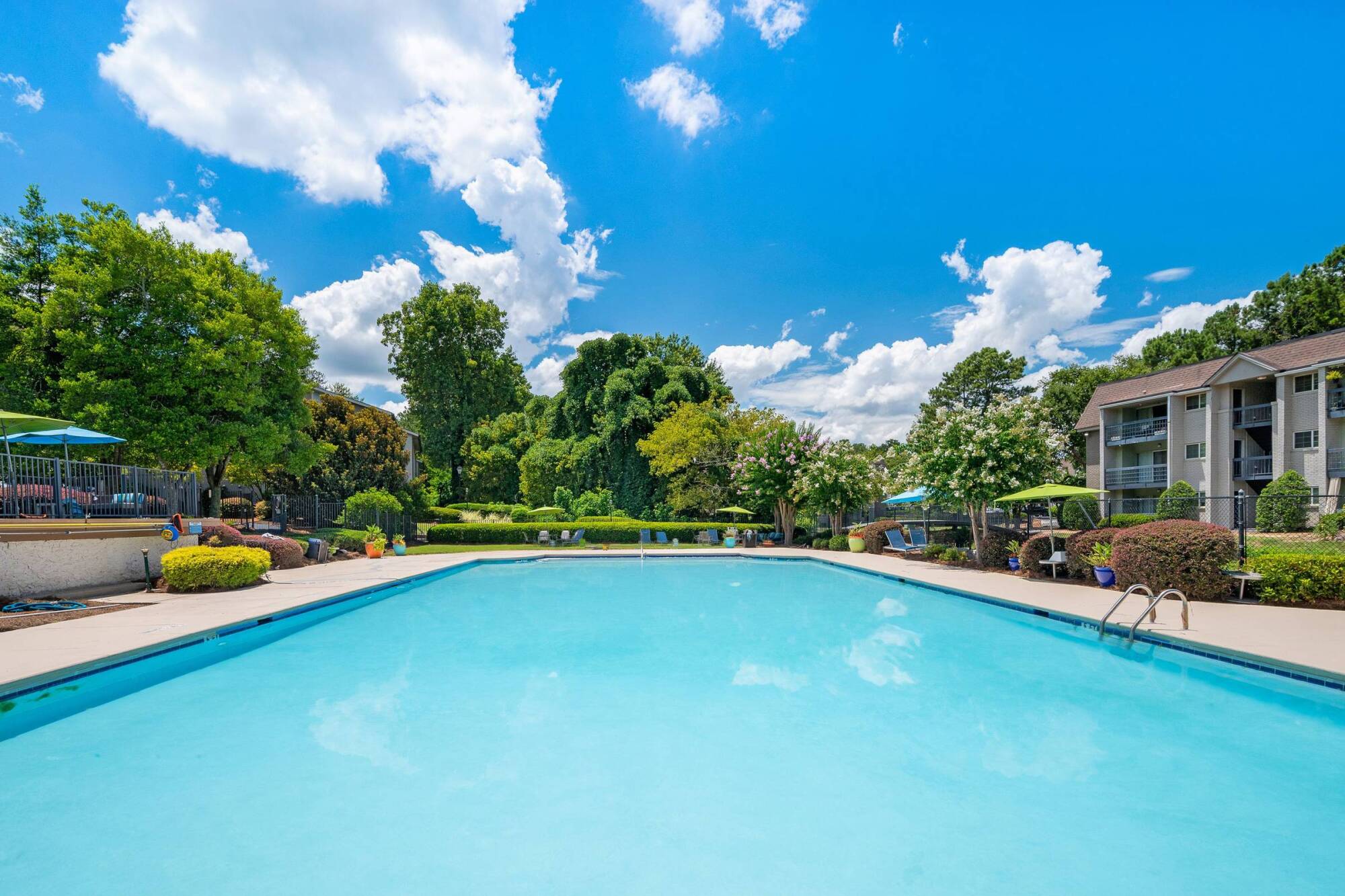 A clear outdoor swimming pool surrounded by trees and lounge chairs under a bright blue sky with clouds.