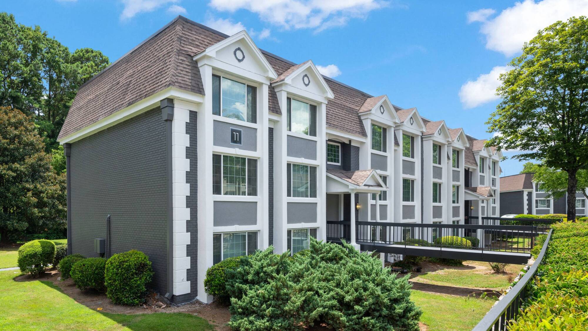 Three-story modern apartment building with gray walls, white trim, large windows, and shrubs in front.