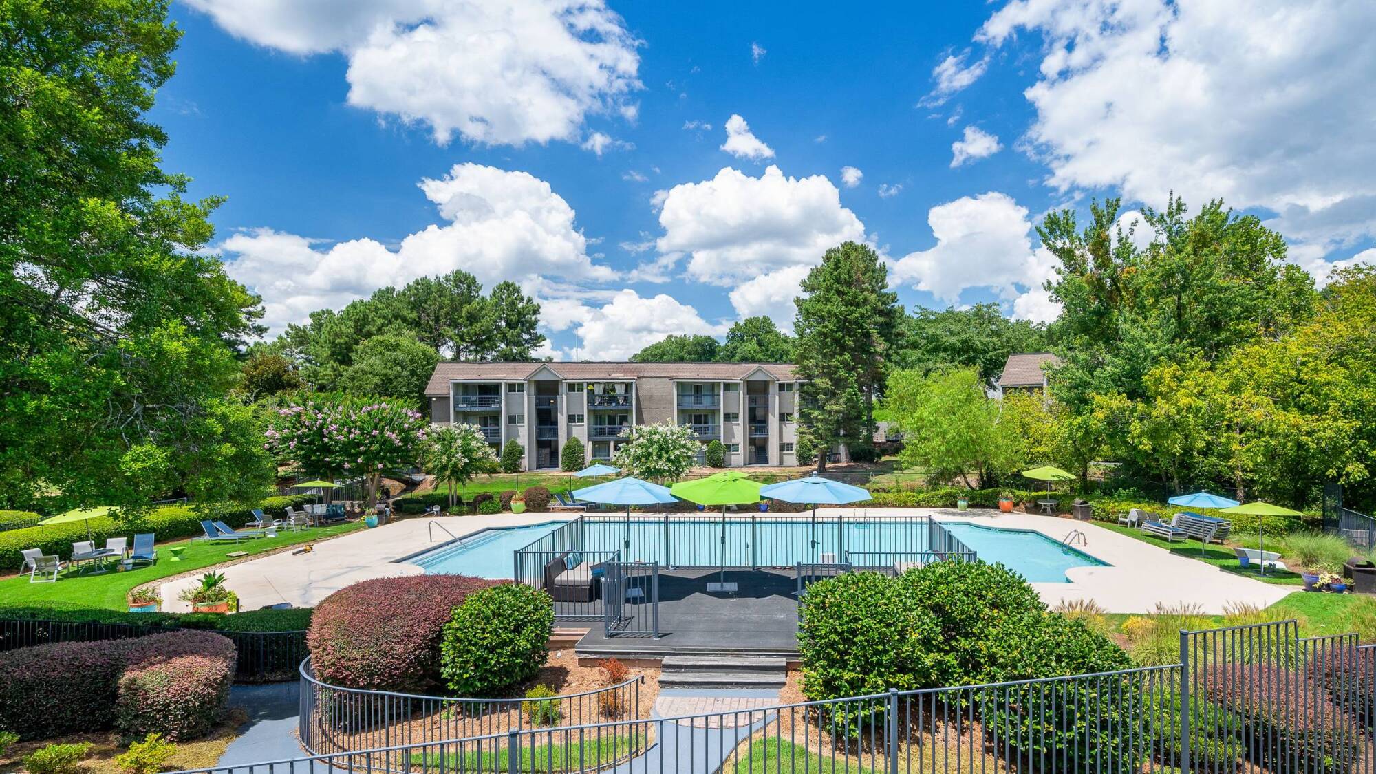 Apartment complex with outdoor pool, green trees, and blue sky with clouds in the background.