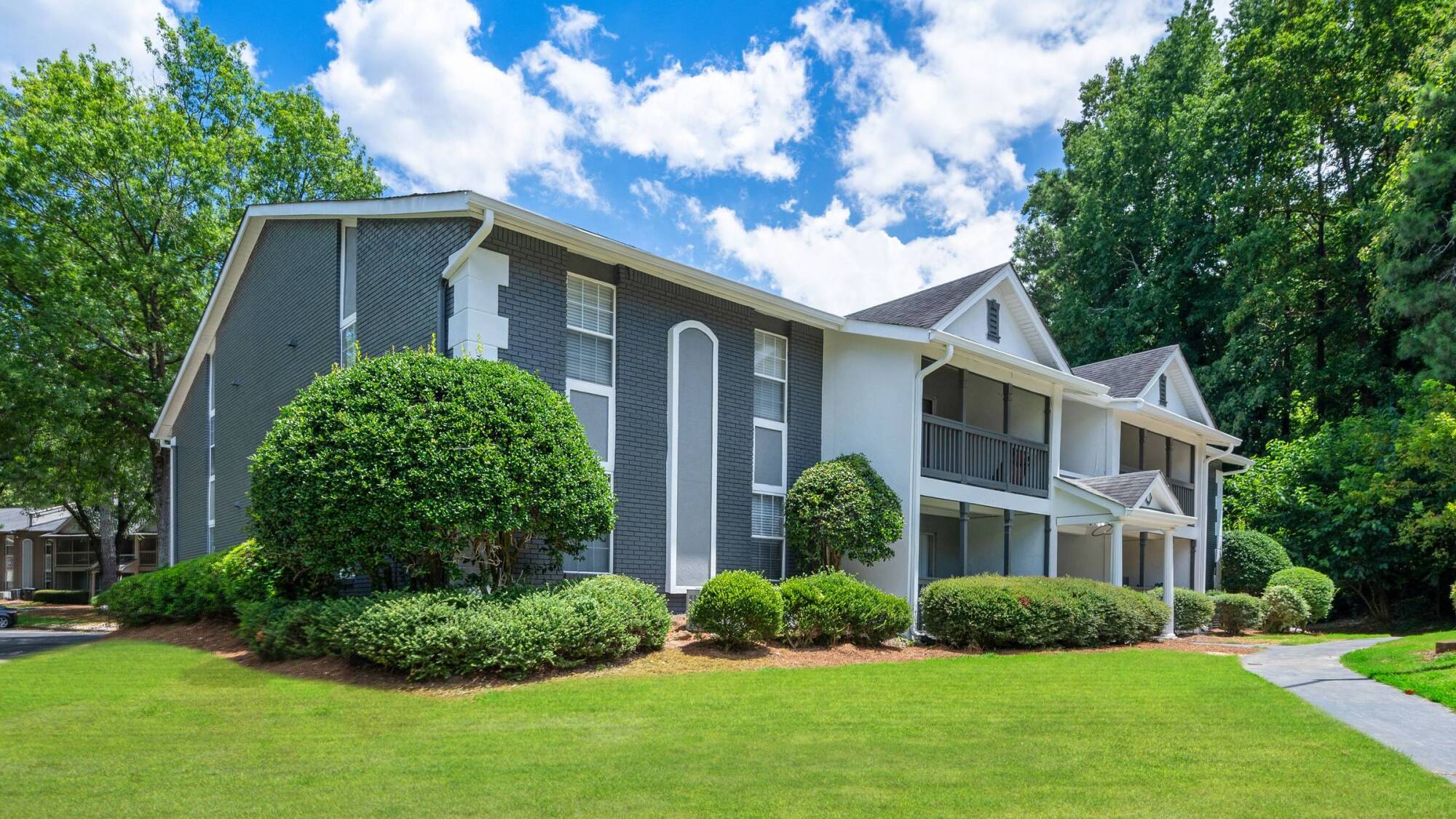Two-story gray and white apartment building with balconies, surrounded by green lawn and bushes.