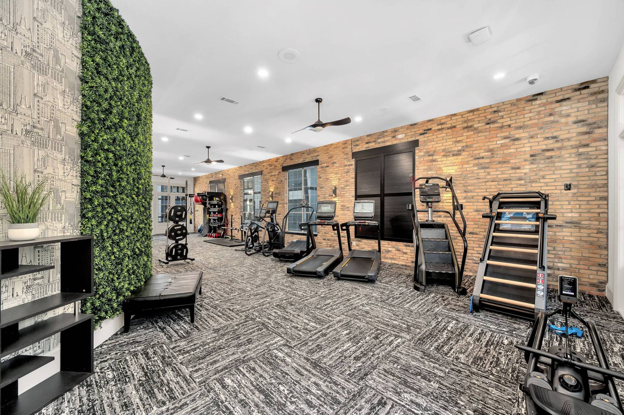 Modern gym with brick walls, treadmills, elliptical machines, weights, and a green wall feature.