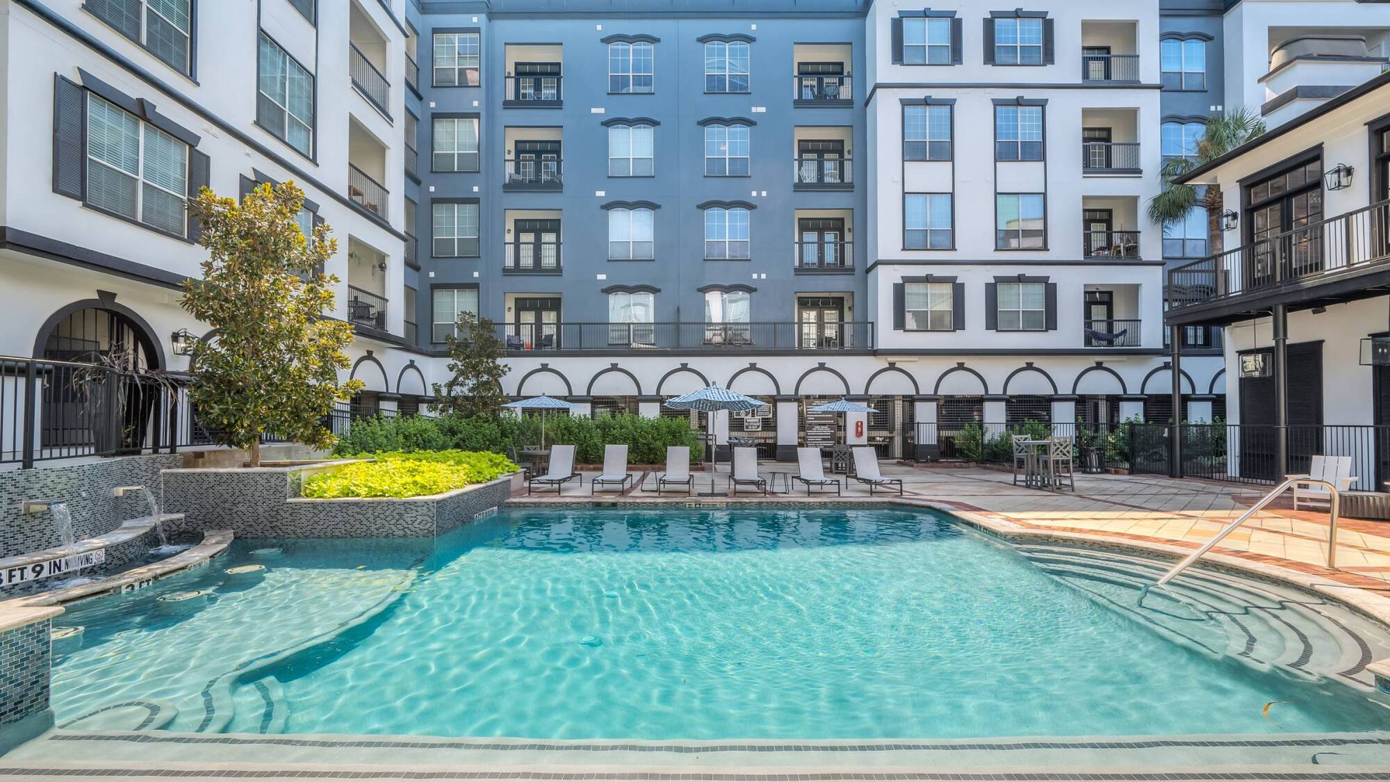 Rectangular outdoor pool with lounge chairs, greenery, and a multi-story apartment building in the background.