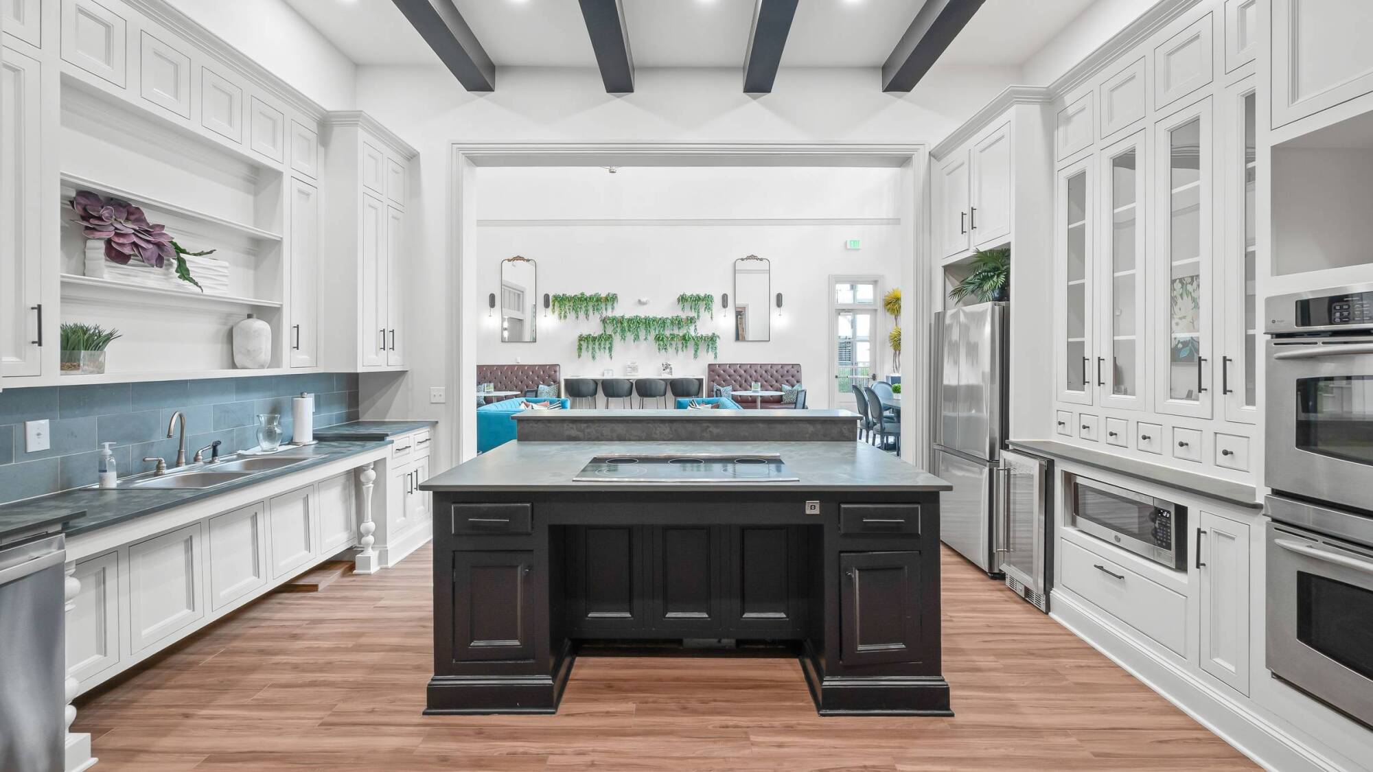 Modern kitchen with black island, white cabinets, wood floors, and dining area with green plants in the background.