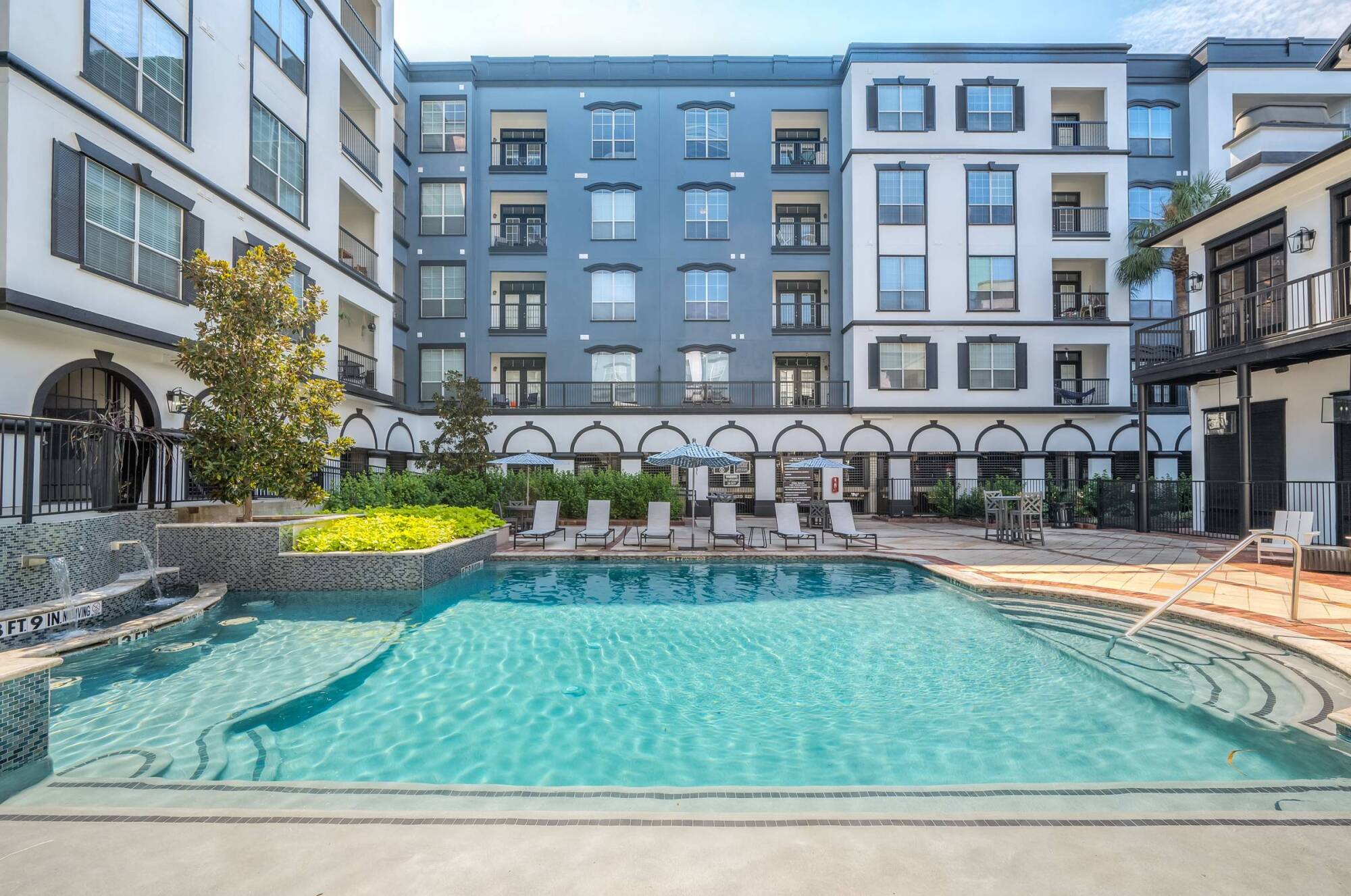 Outdoor swimming pool with lounge chairs in front of a modern apartment building on a sunny day.