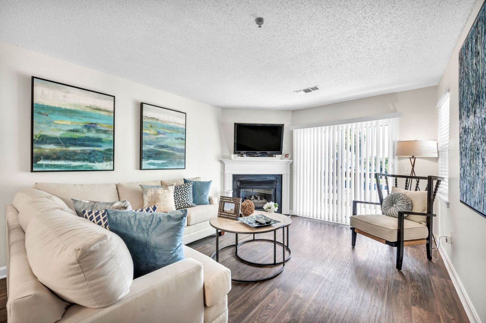Bright living room with a cream sofa, art on the wall, fireplace, TV, armchair, and sliding glass doors.