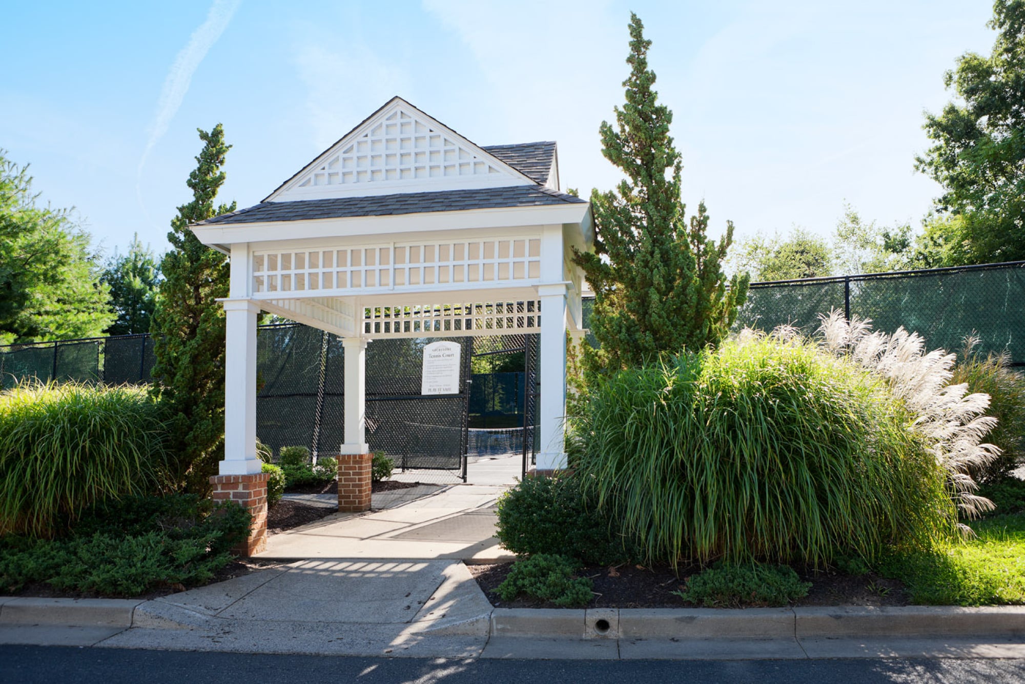 Milestone Apartments White gazebo entrance surrounded by greenery, leading to tennis courts on a sunny day.