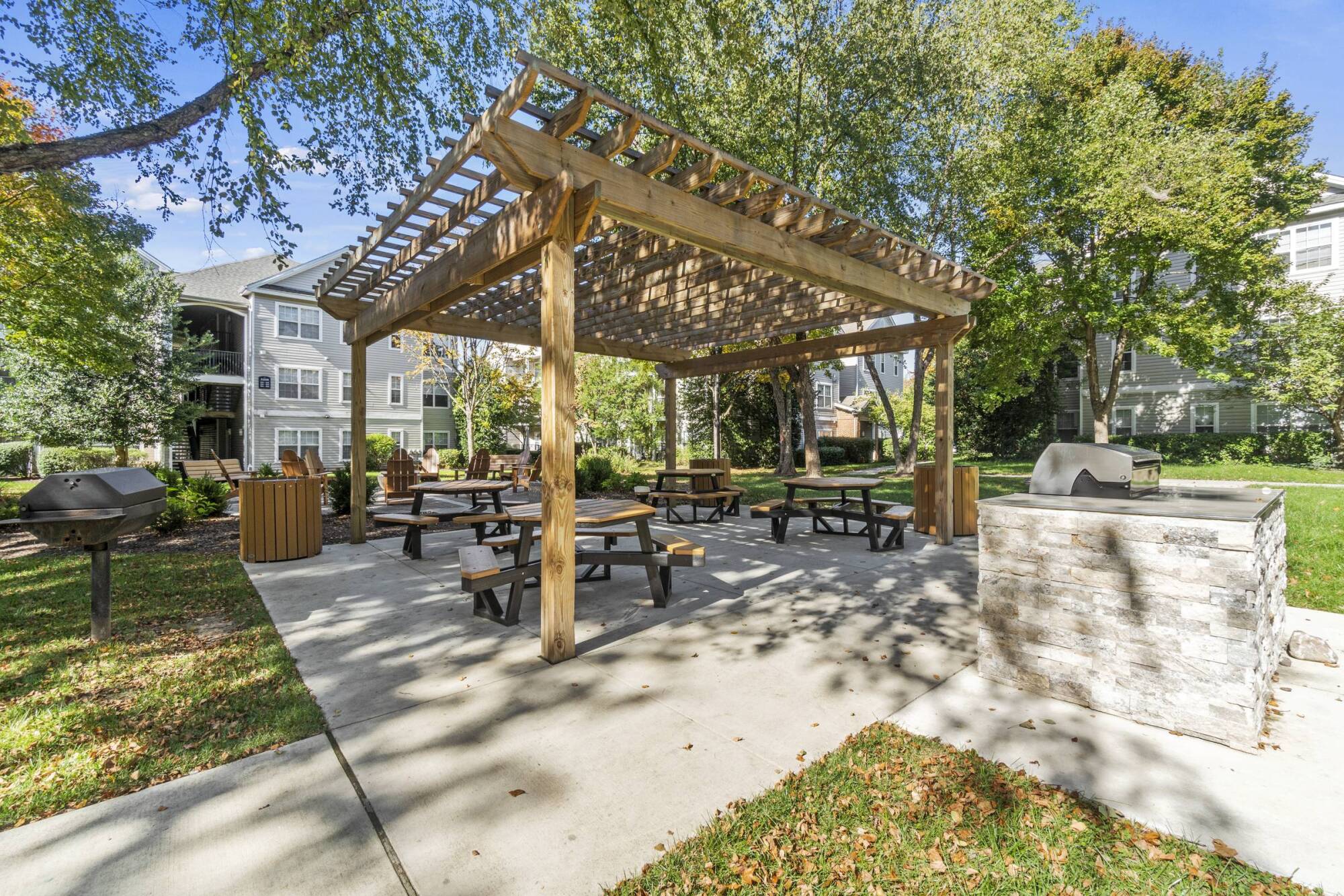 Milestone Apartments Wood pergola with picnic tables and grills in a sunny outdoor apartment courtyard surrounded by trees.