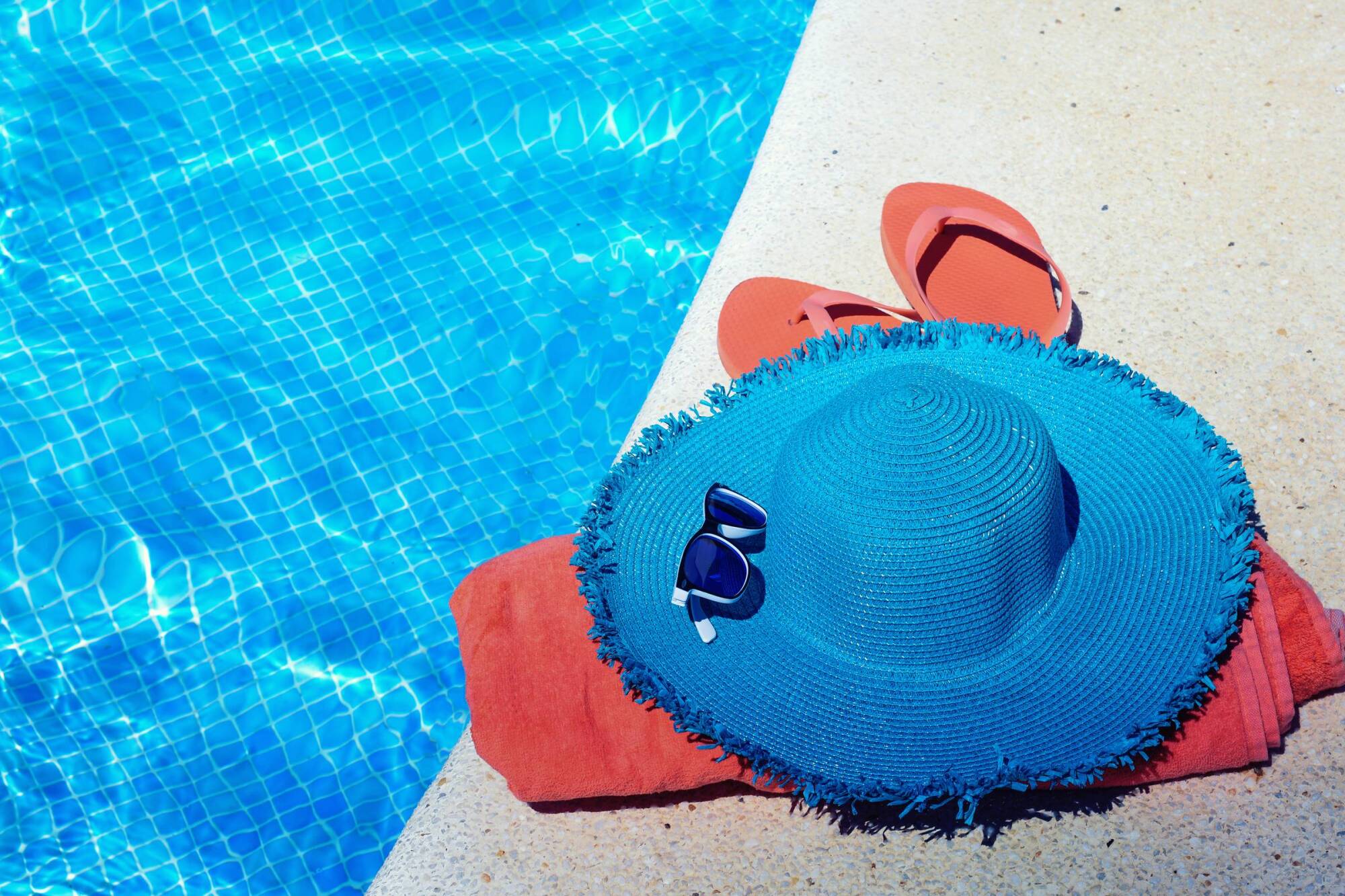 Blue sun hat, sunglasses, flip flops, and towel by blue pool
