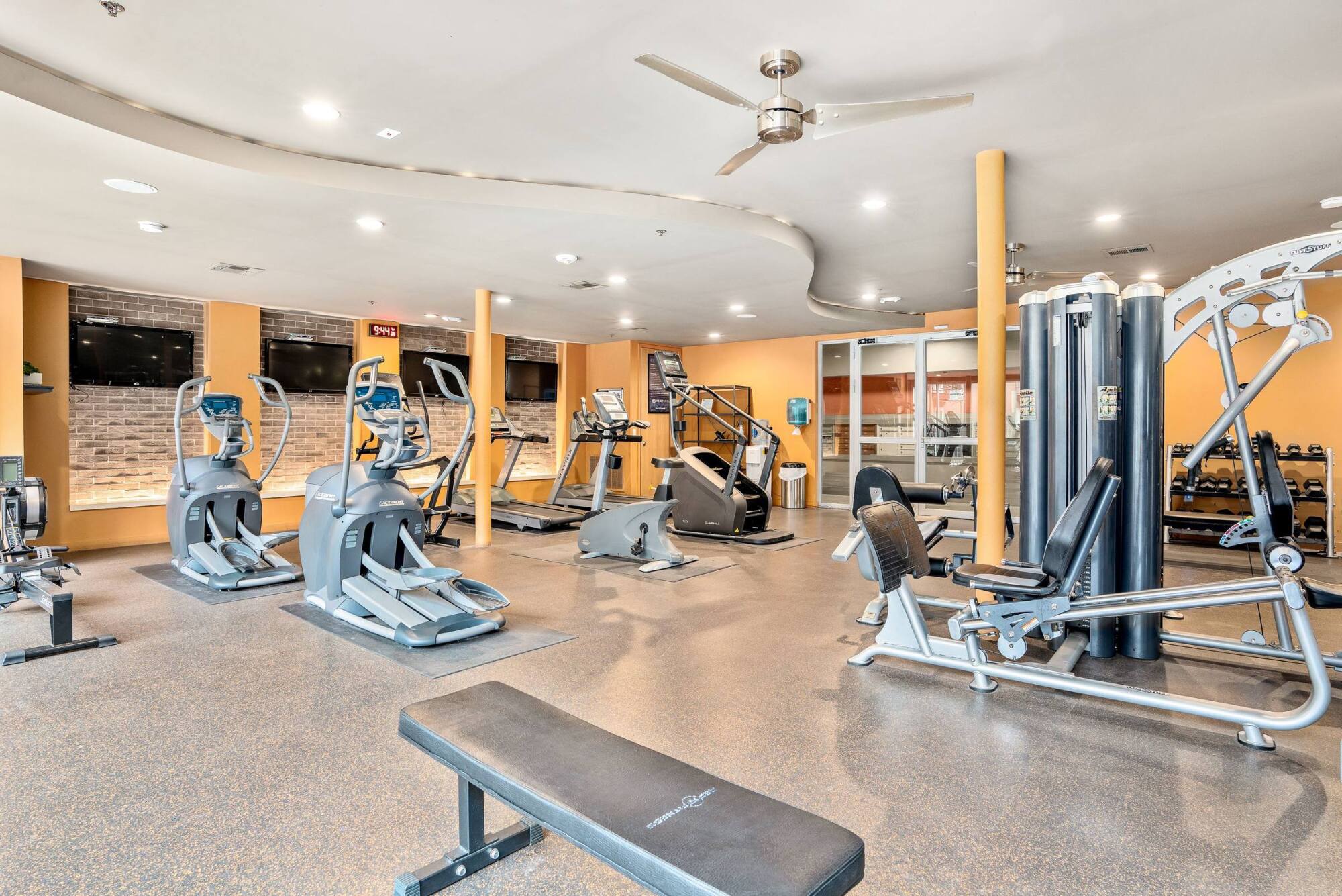 Modern gym with cardio machines, weight equipment, benches, and TVs in a bright, spacious room.