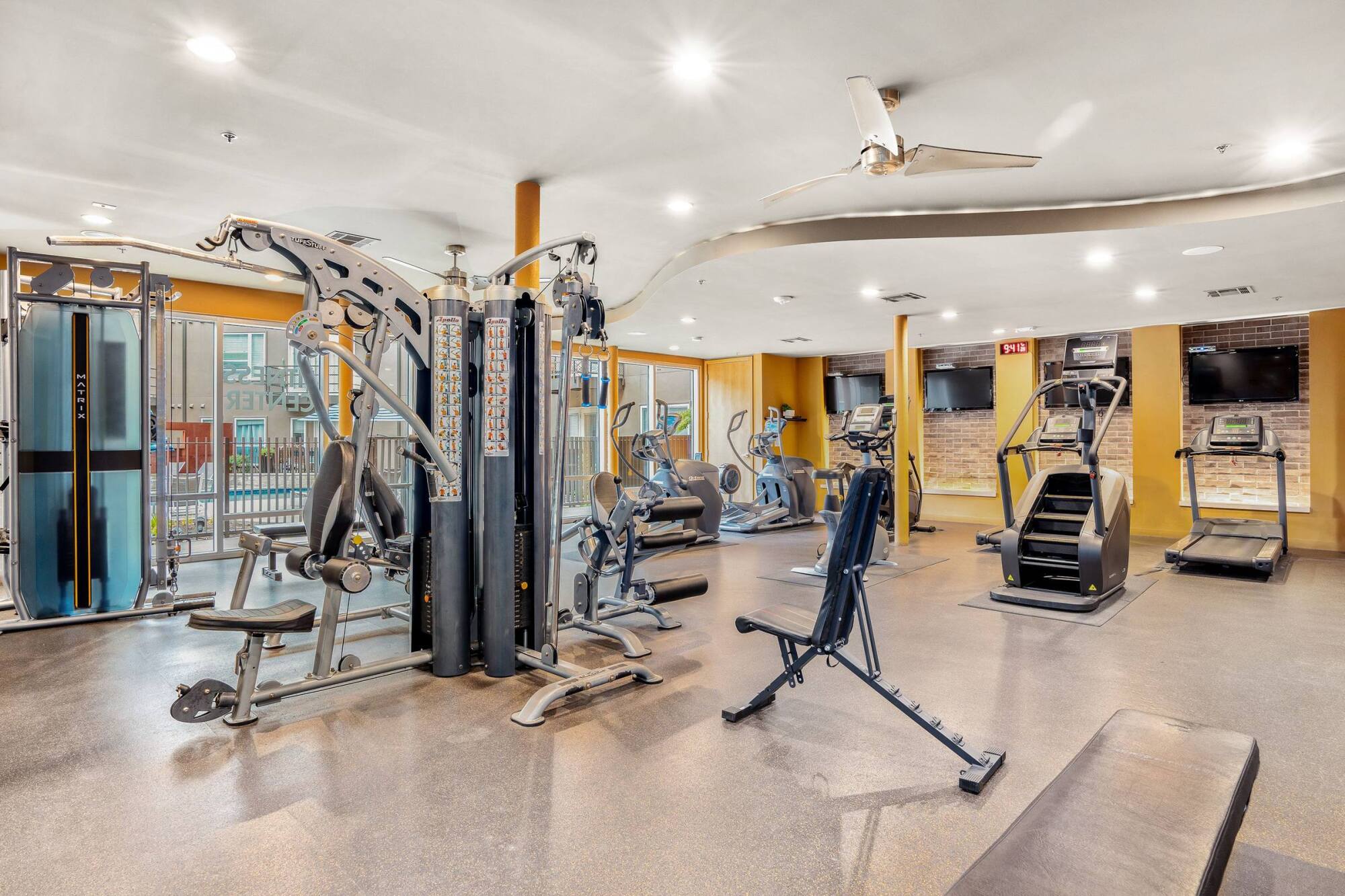 Modern gym with strength and cardio machines, benches, and large windows letting in natural light.