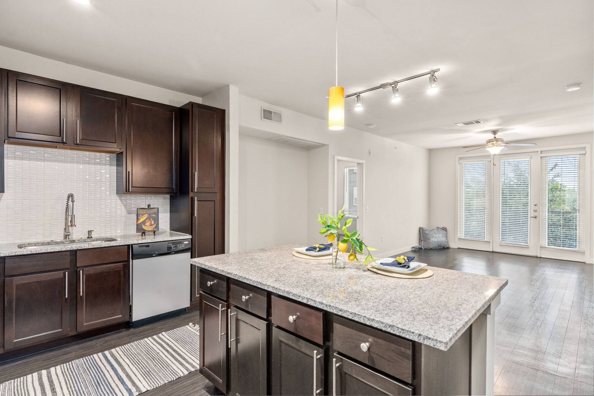 Modern kitchen with dark cabinets, granite island, pendant light, and open living area with large windows.