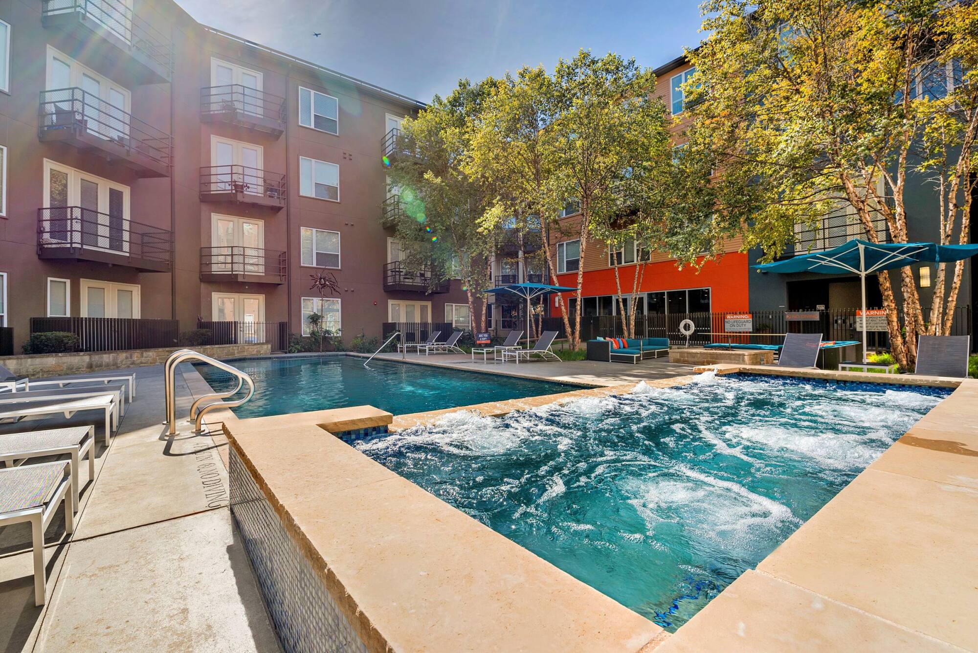 Outdoor pool and hot tub area with lounge chairs, umbrellas, and apartment buildings in the background.