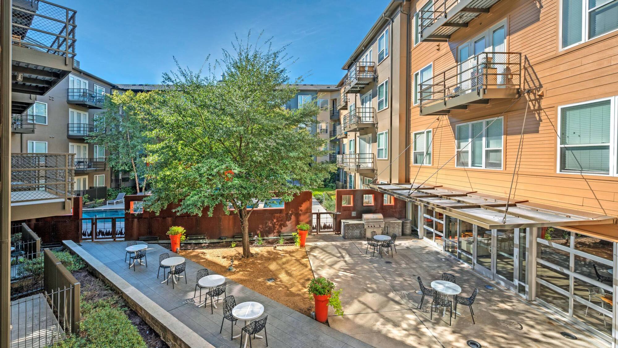 Apartment courtyard with small tables, chairs, potted plants, and a tree surrounded by multi-story buildings.