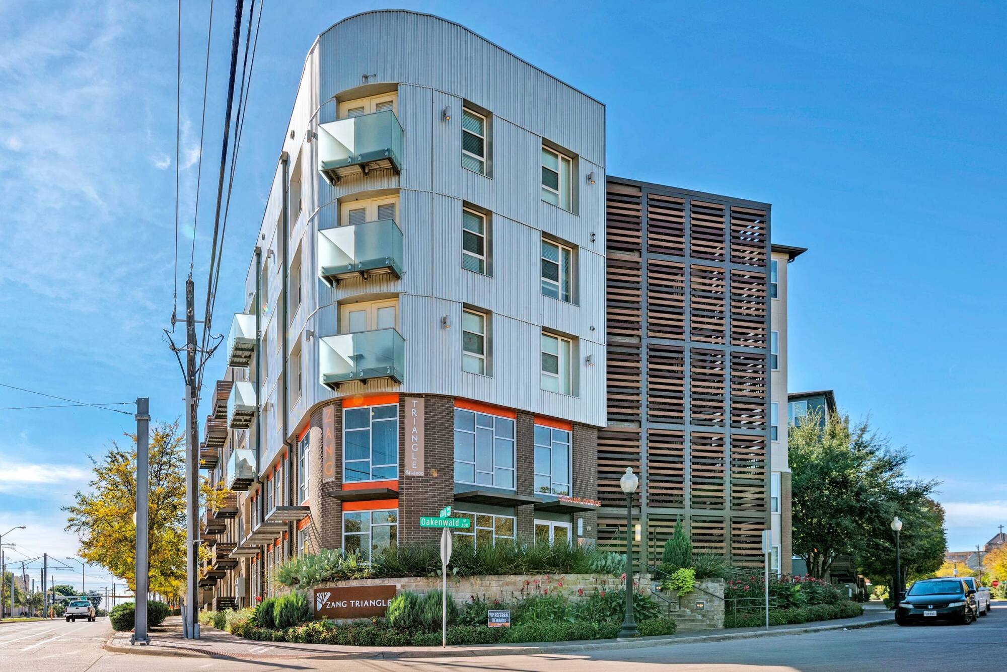 Modern four-story apartment building with balconies on a sunny corner street, surrounded by greenery.