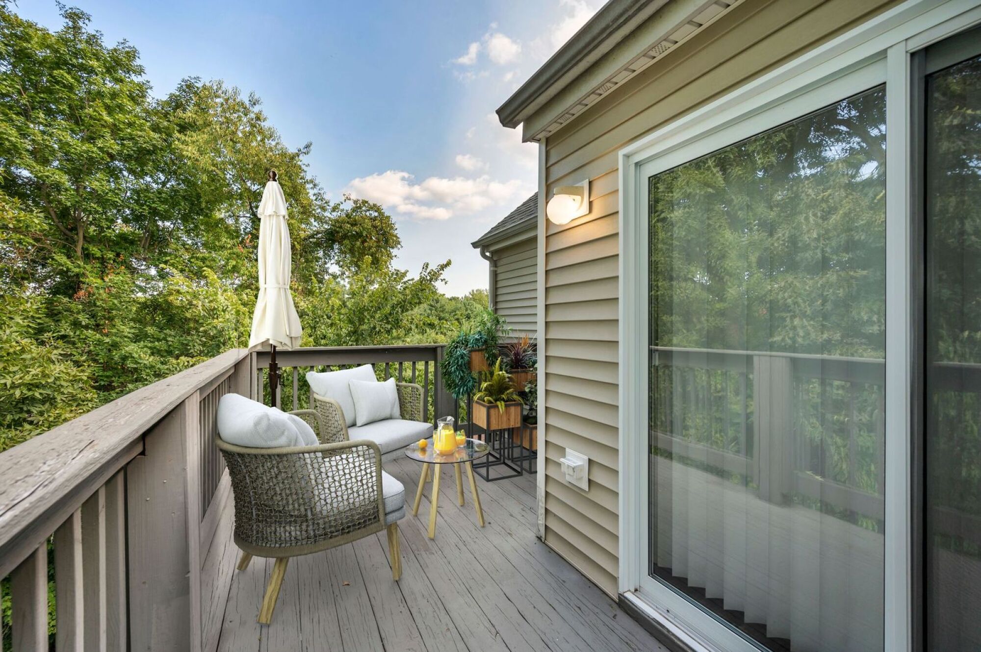 Small balcony with two chairs, a side table, potted plants, and a leafy view beside a sliding glass door.