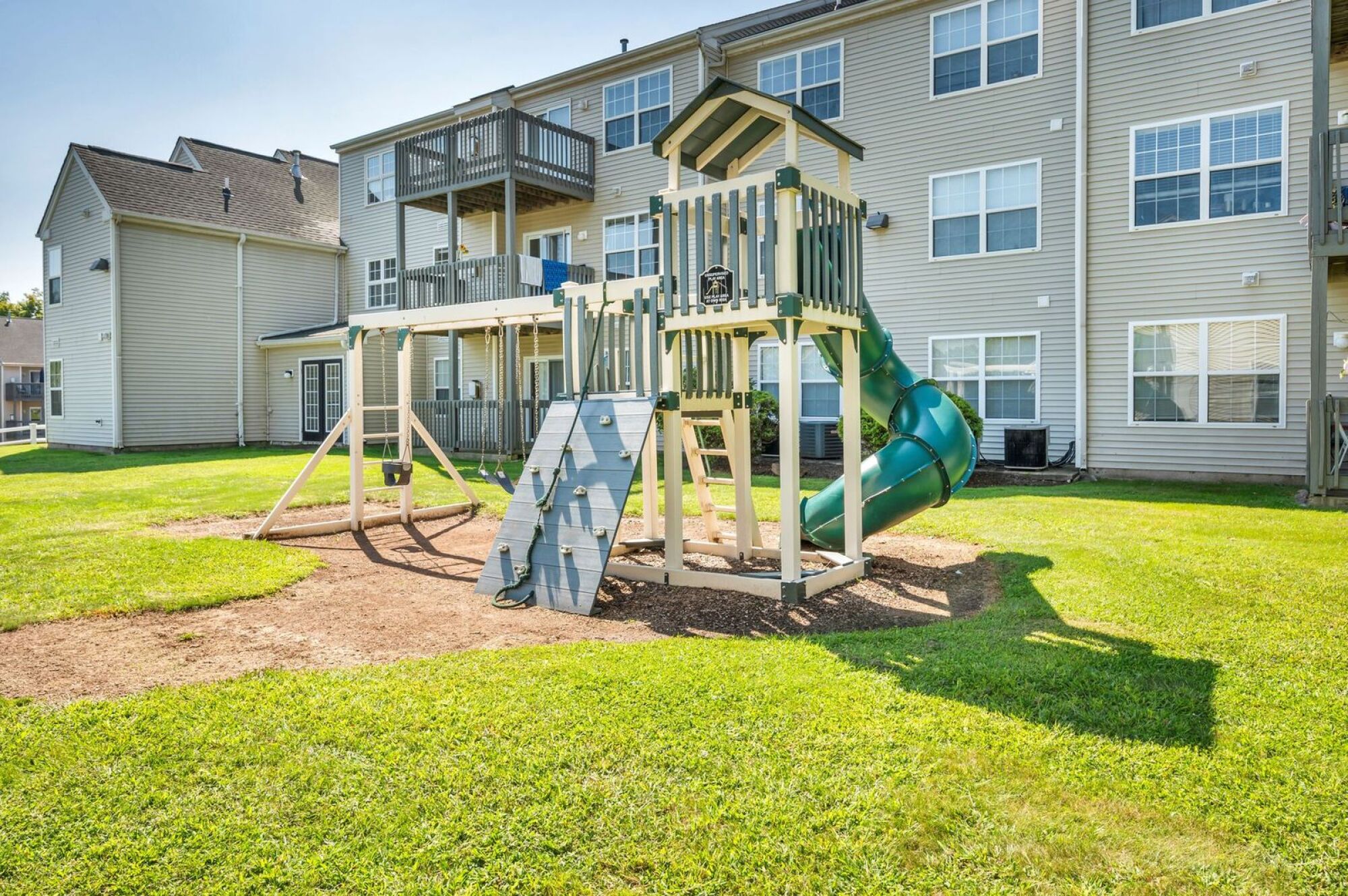 Playground with swings, climbing wall, and green spiral slide in front of an apartment building on a sunny day.