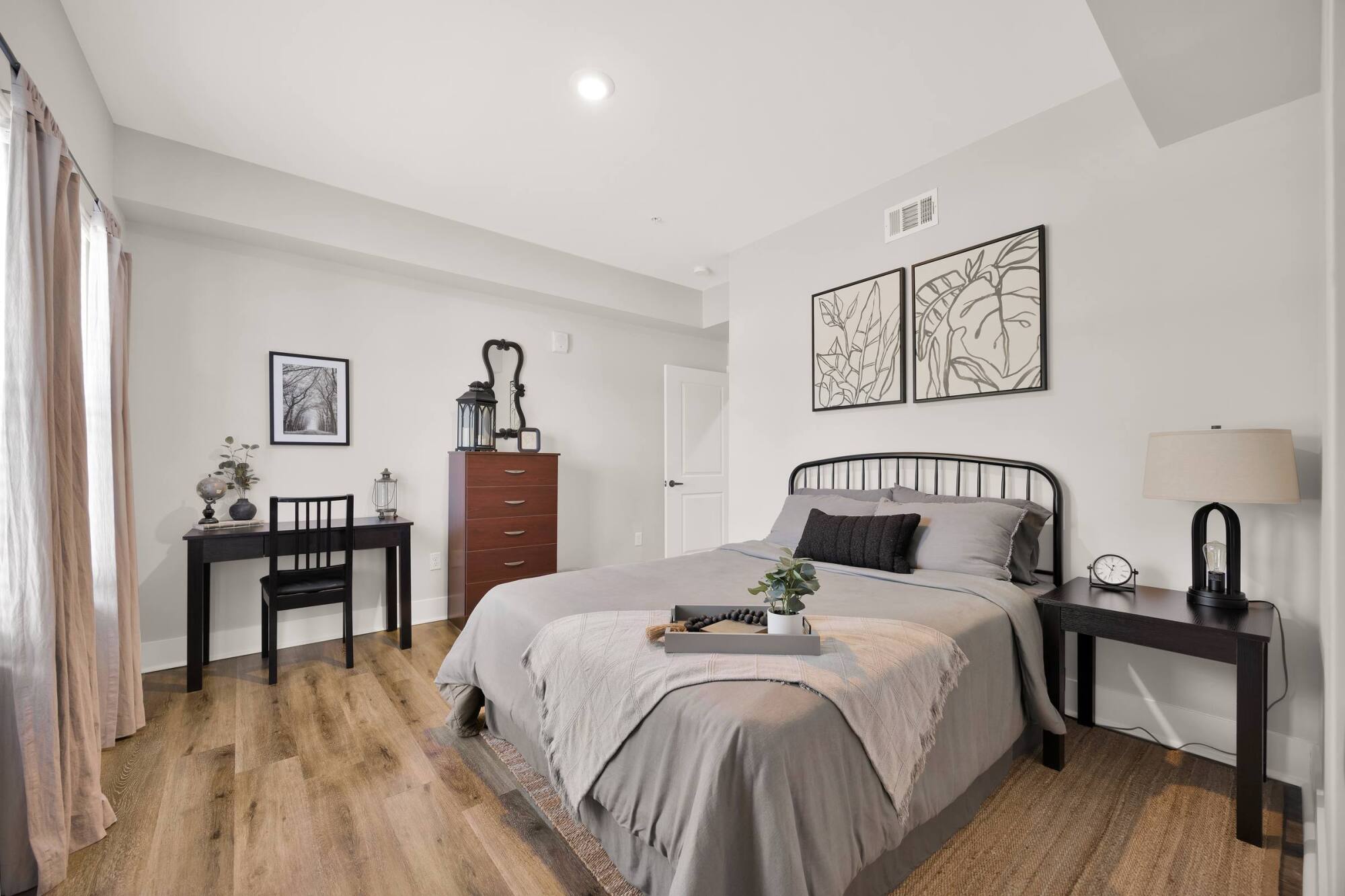 A modern bedroom with a black bed, gray bedding, desk, chest of drawers, and minimalist wall art.