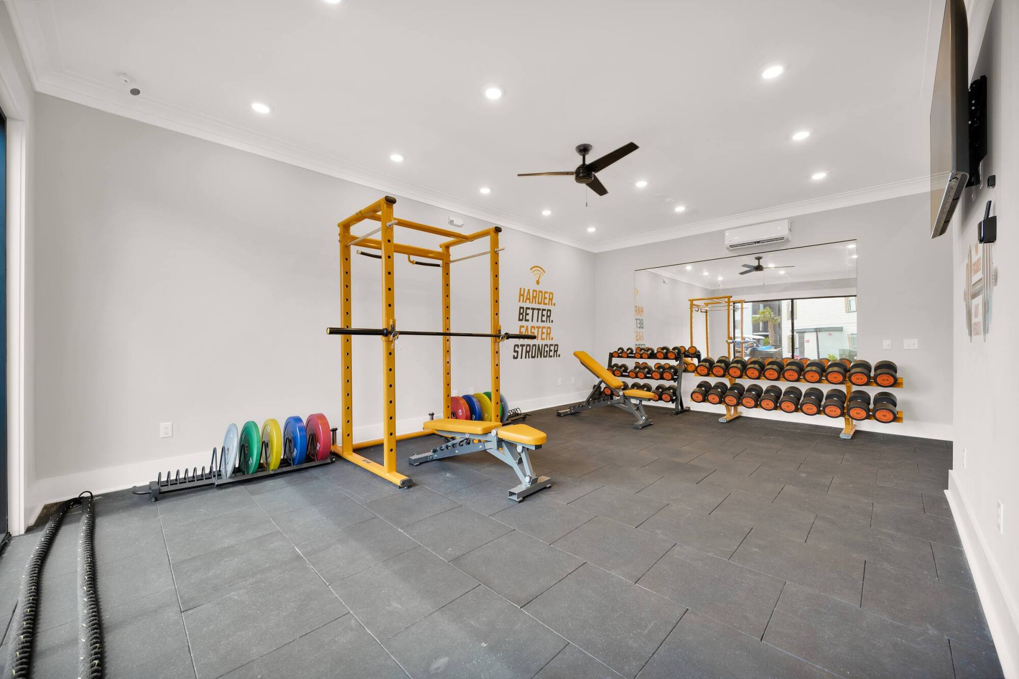 Modern gym with yellow squat rack, weight bench, dumbbells, and motivational quote on the wall.