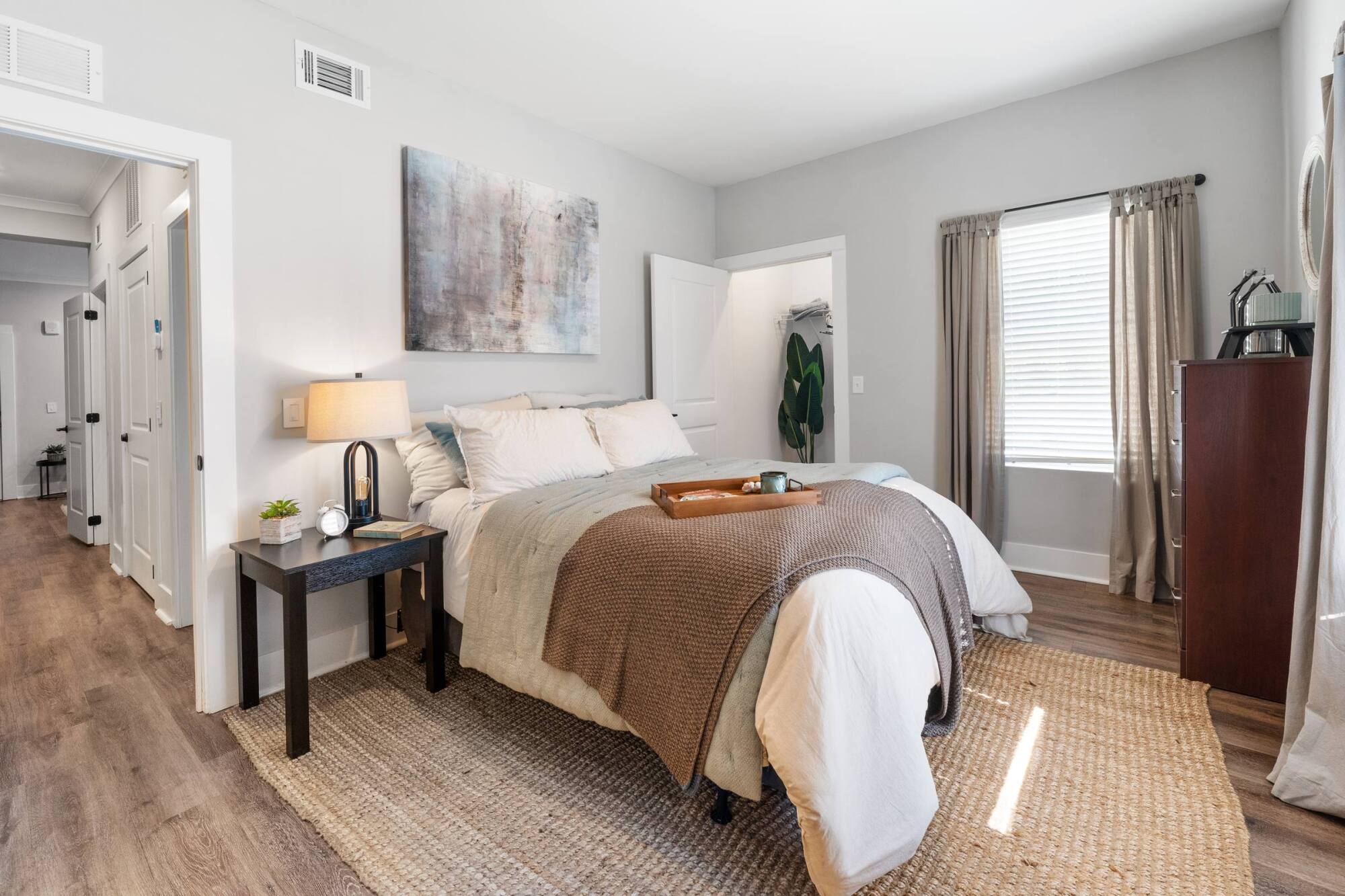 Bright bedroom with a neatly made bed, bedside table, and a window with light curtains letting in sunlight.