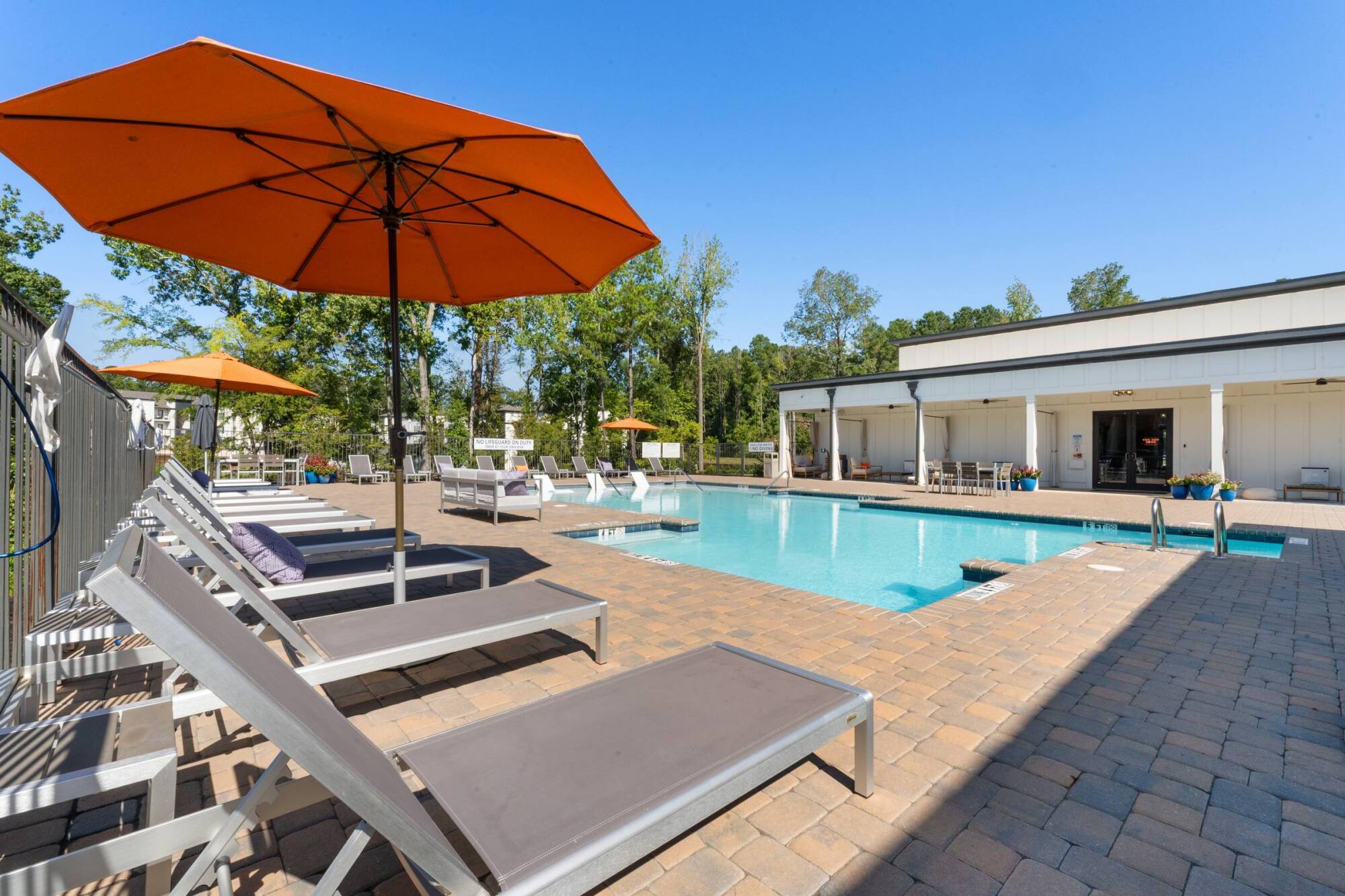Outdoor pool area with lounge chairs, large orange umbrellas, and a white poolside building under a clear sky.