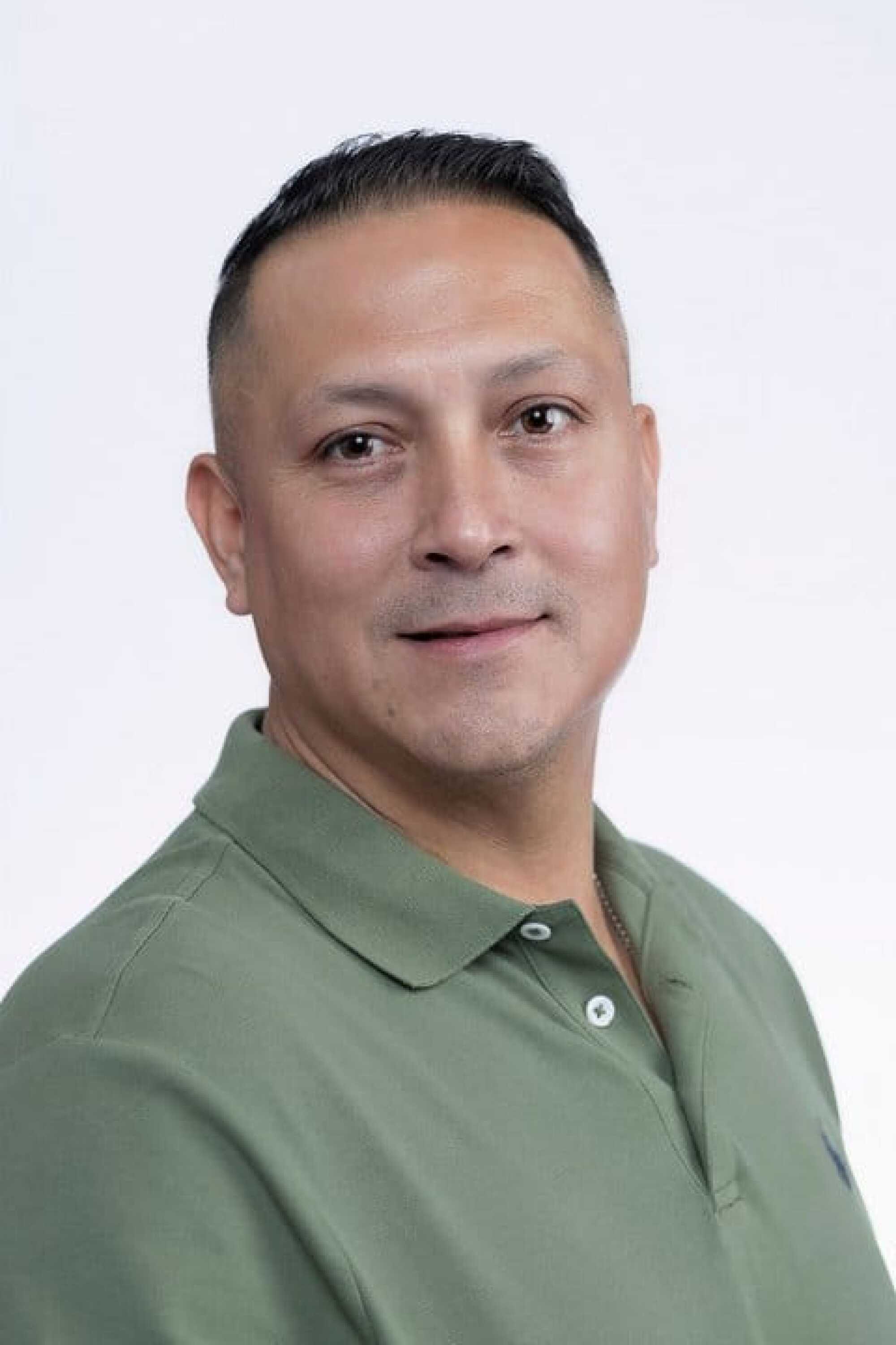 Man with short dark hair wearing a green collared shirt smiles slightly at the camera against a plain light background.