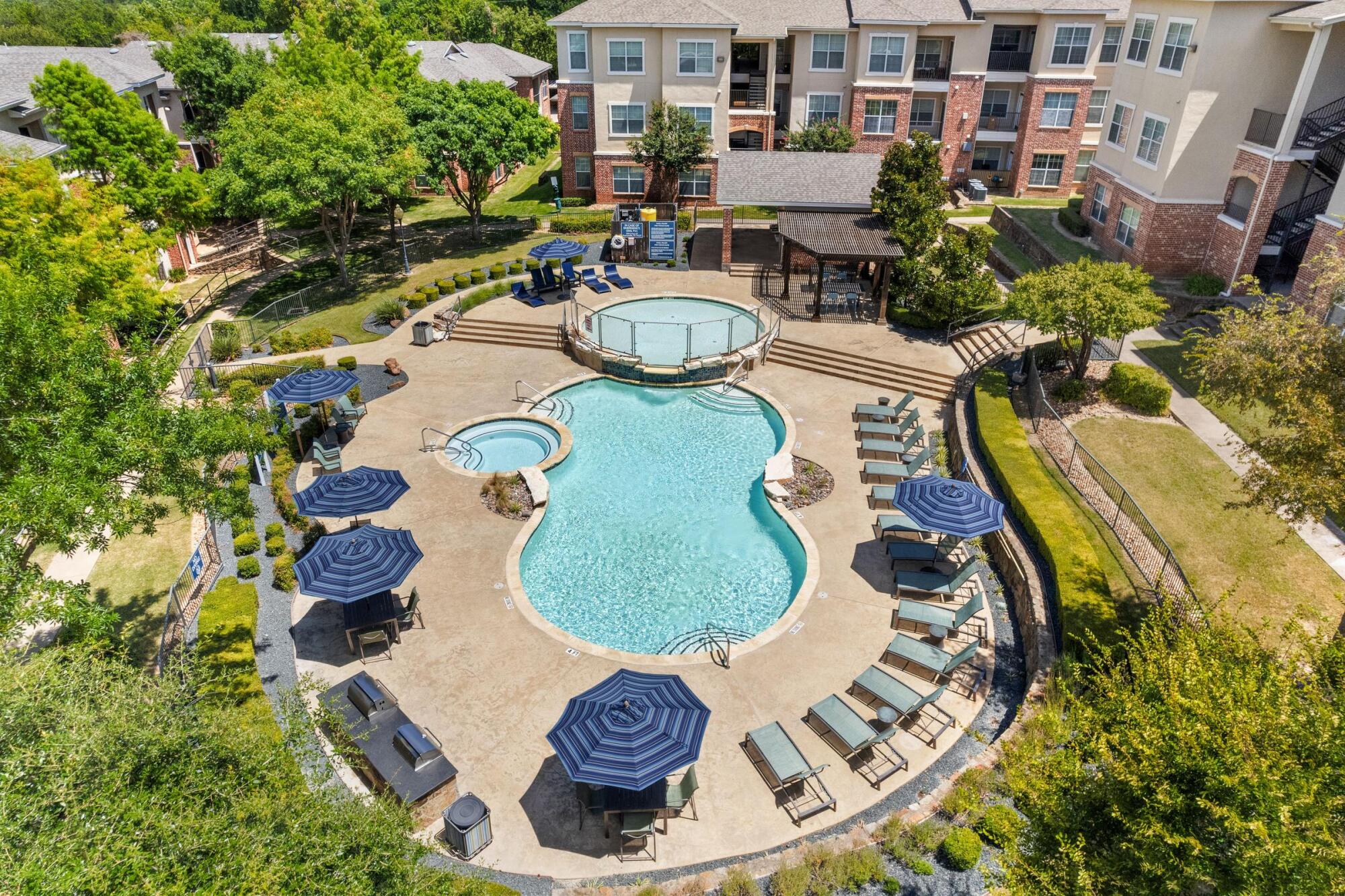 Aerial view of the pool and hot tub at Bella Ruscello in Duncanville, TX with blue umbrellas, lounge chairs, and landscaped grounds