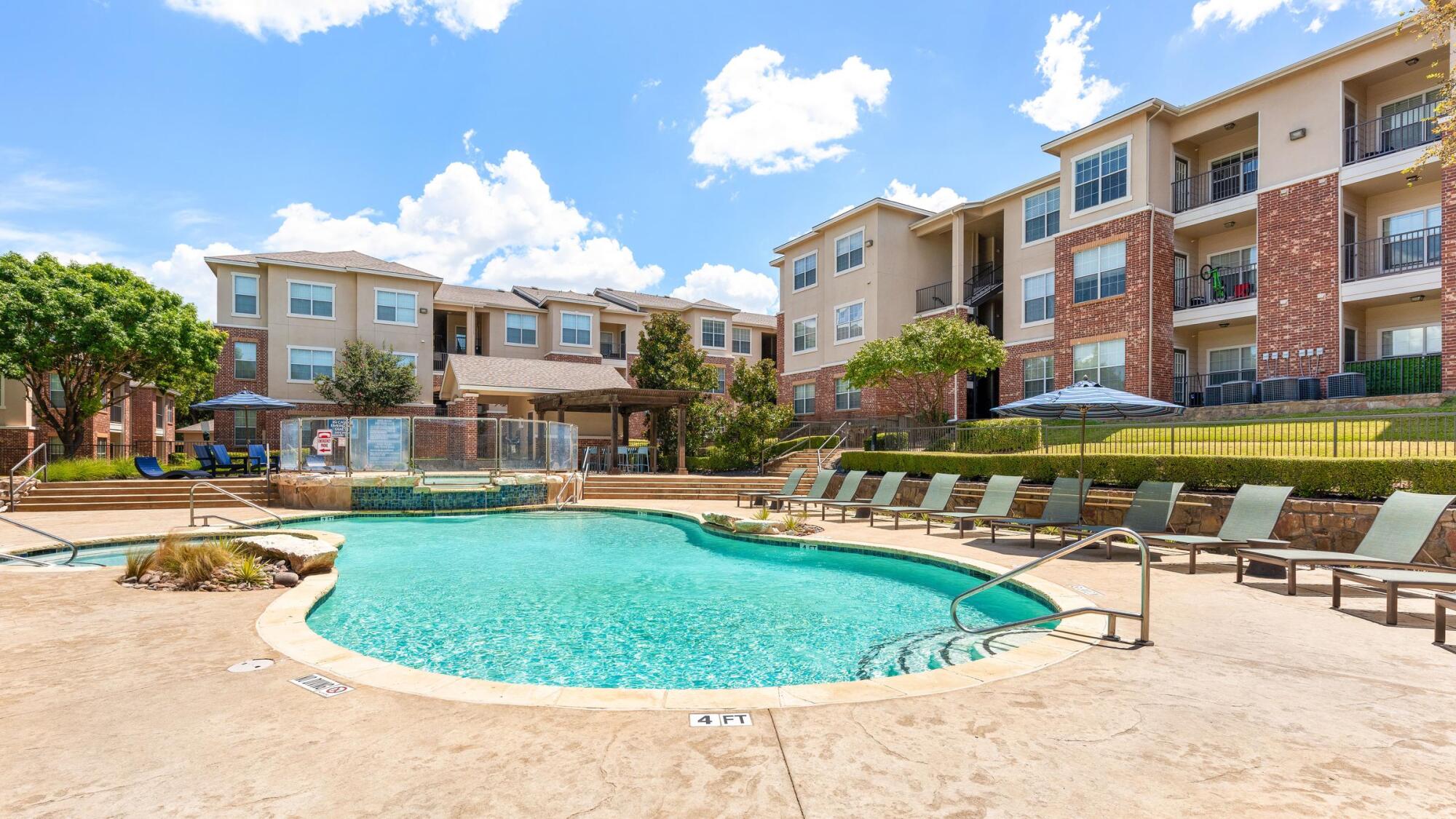 Resort-style swimming pool at Bella Ruscello in Duncanville, TX with lounge chairs, covered pavilion, and brick apartment buildings