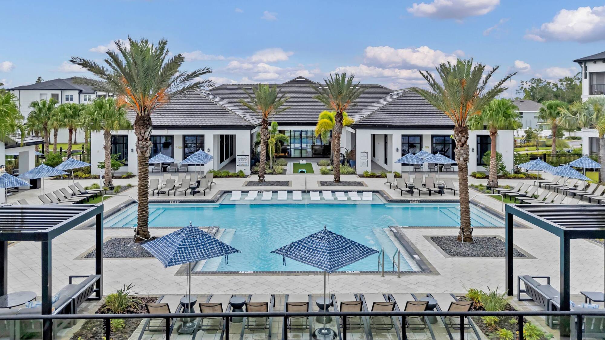 Resort-style pool with lounge chairs, palm trees, umbrellas, and modern buildings in the background.