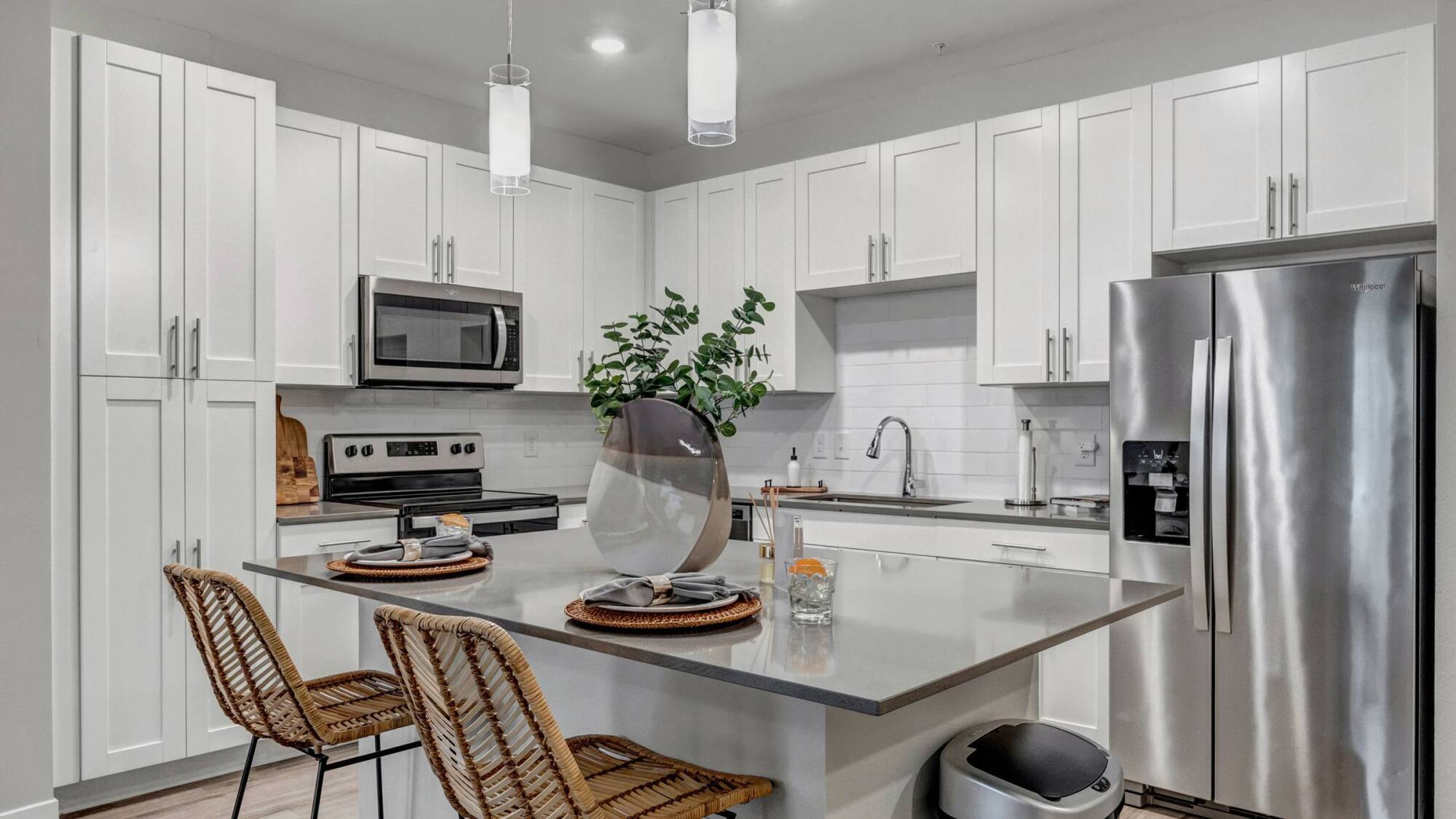 Modern white kitchen with stainless steel appliances, island seating, and decorative plants on the counter.