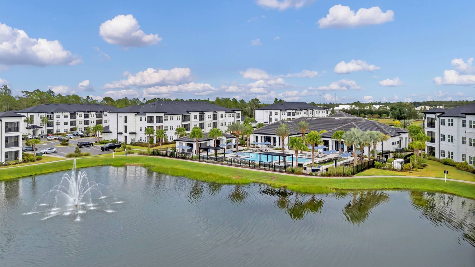 A modern apartment complex with a pool, palm trees, and a lake with a fountain under a blue sky.