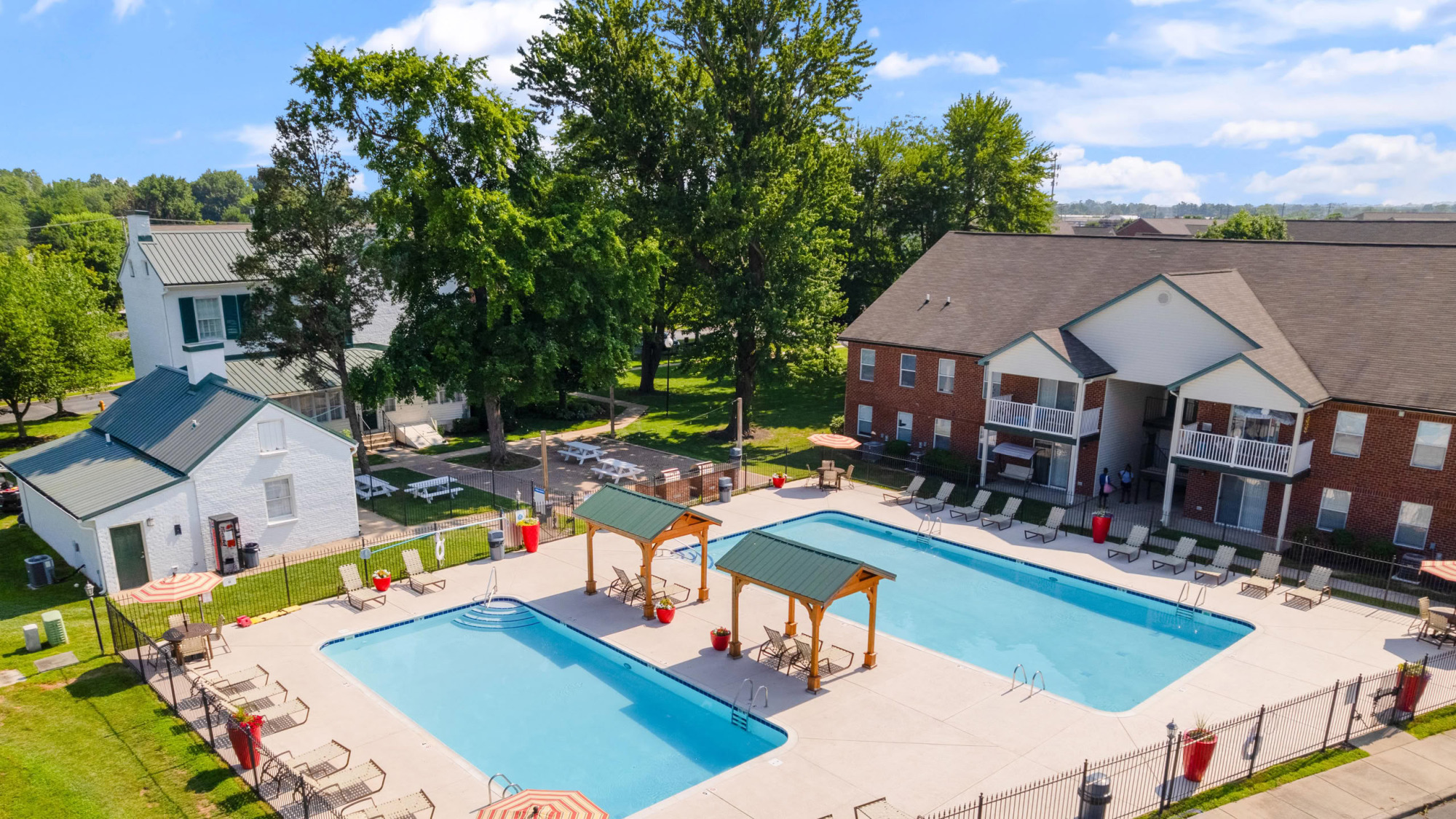 Aerial view of a brick apartment community featuring a large rectangular swimming pool with poolside lounge chairs, wooden pergolas, mature trees, and surrounding residential buildings.