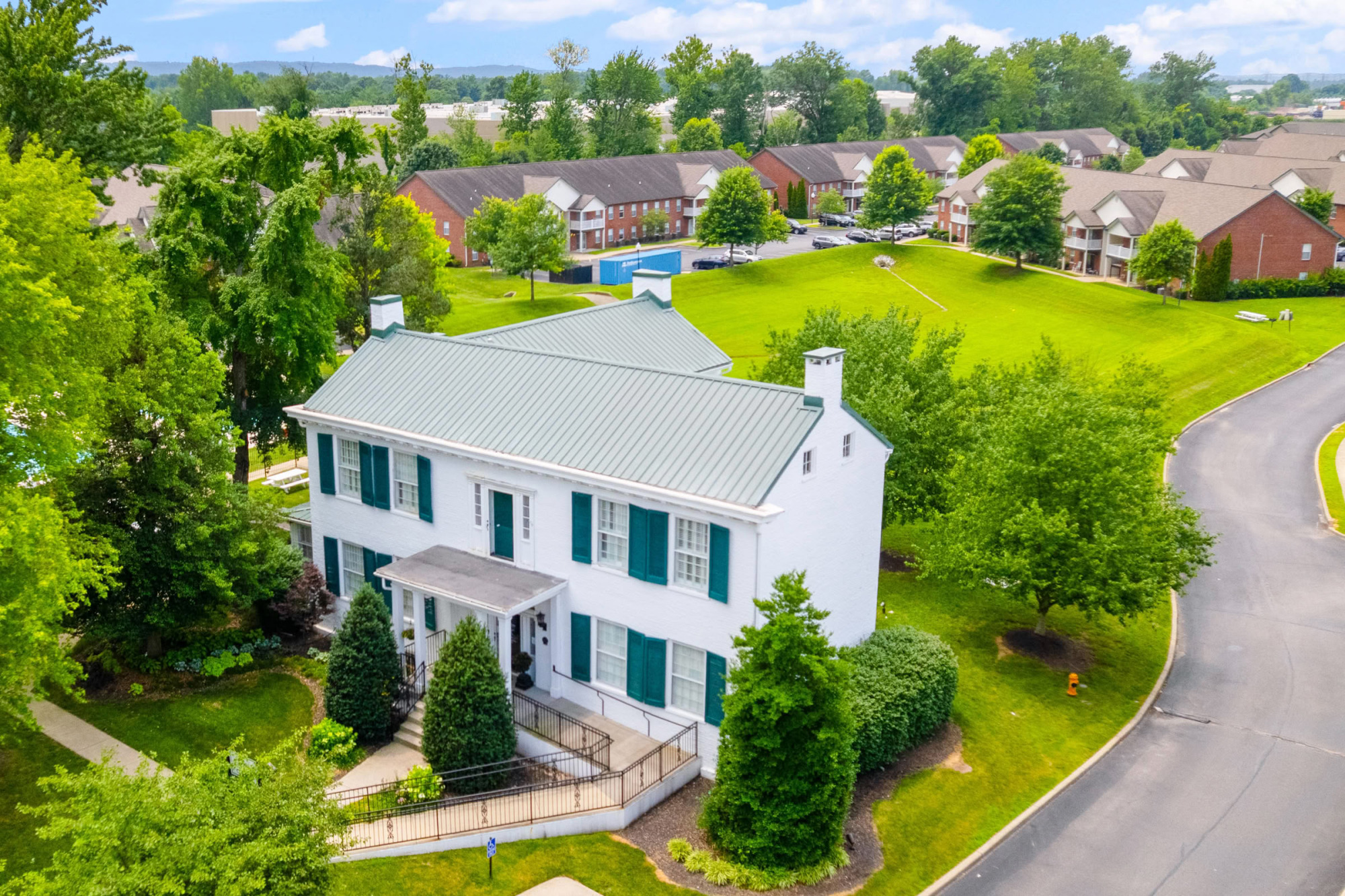 Aerial view of a stately white colonial-style leasing office with a green metal roof and dark shutters, surrounded by manicured lawns, mature trees, and the apartment community in the background.