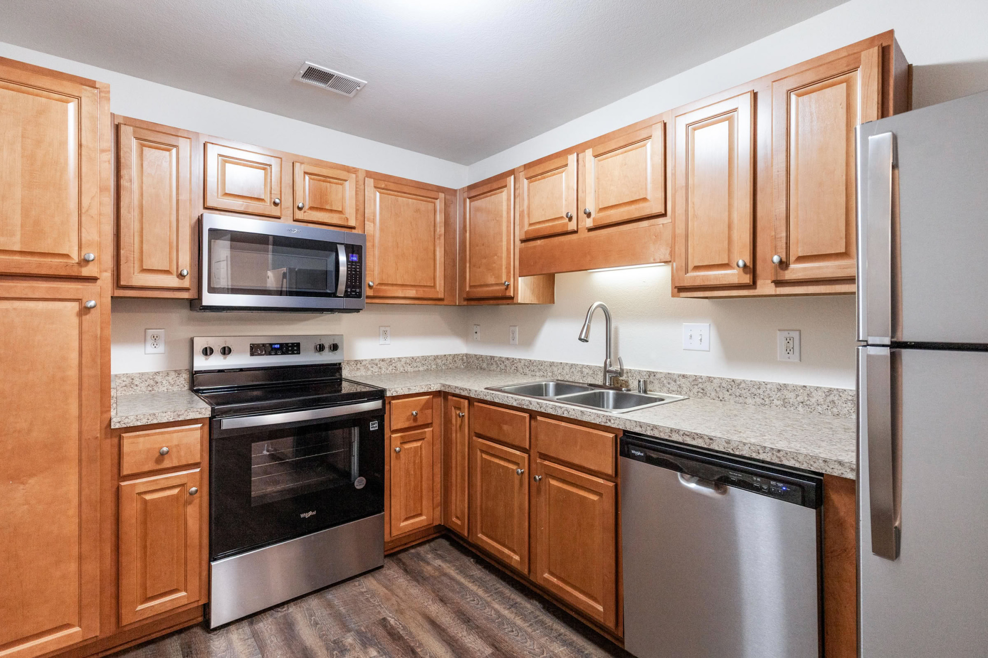 Bright apartment kitchen with maple wood cabinets, stainless steel appliances, granite countertops, and dark hardwood flooring in an L-shaped layout.