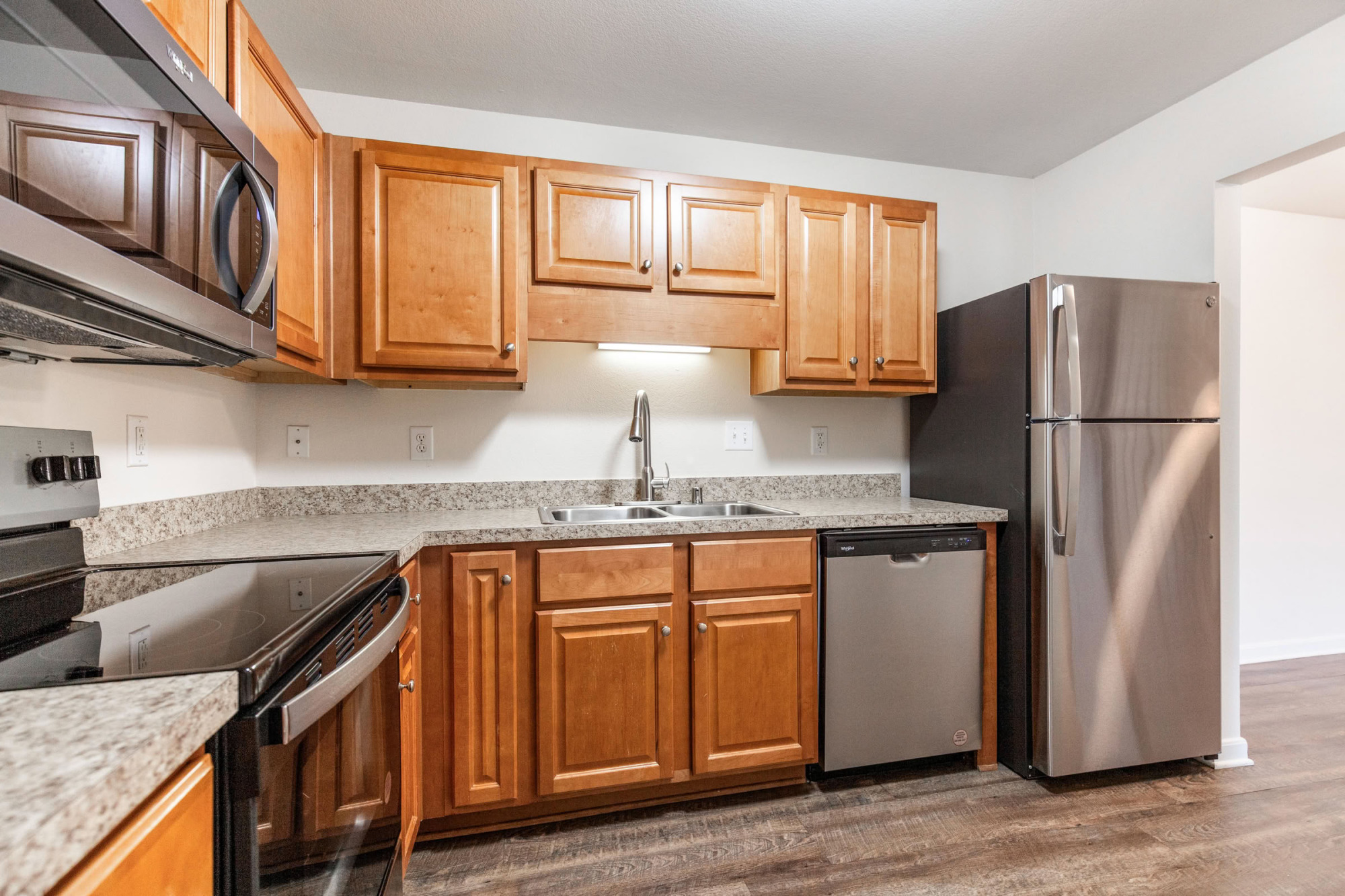 Spacious apartment kitchen featuring honey-toned cabinets, stainless steel refrigerator and dishwasher, granite-style countertops, undercabinet lighting, and rich wood-look flooring.
