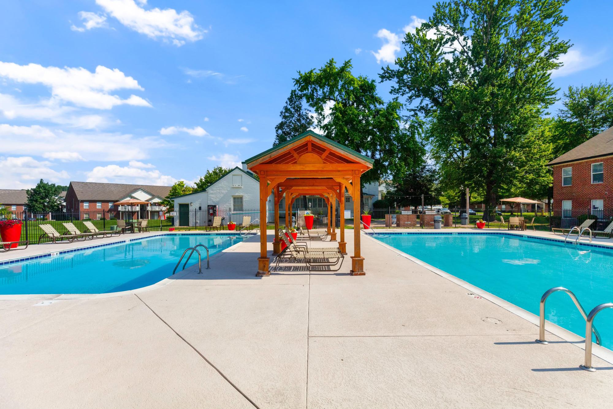 Outdoor swimming pool area featuring a wooden gazebo with shaded lounge chairs, surrounded by green lawn, mature trees, and apartment buildings.