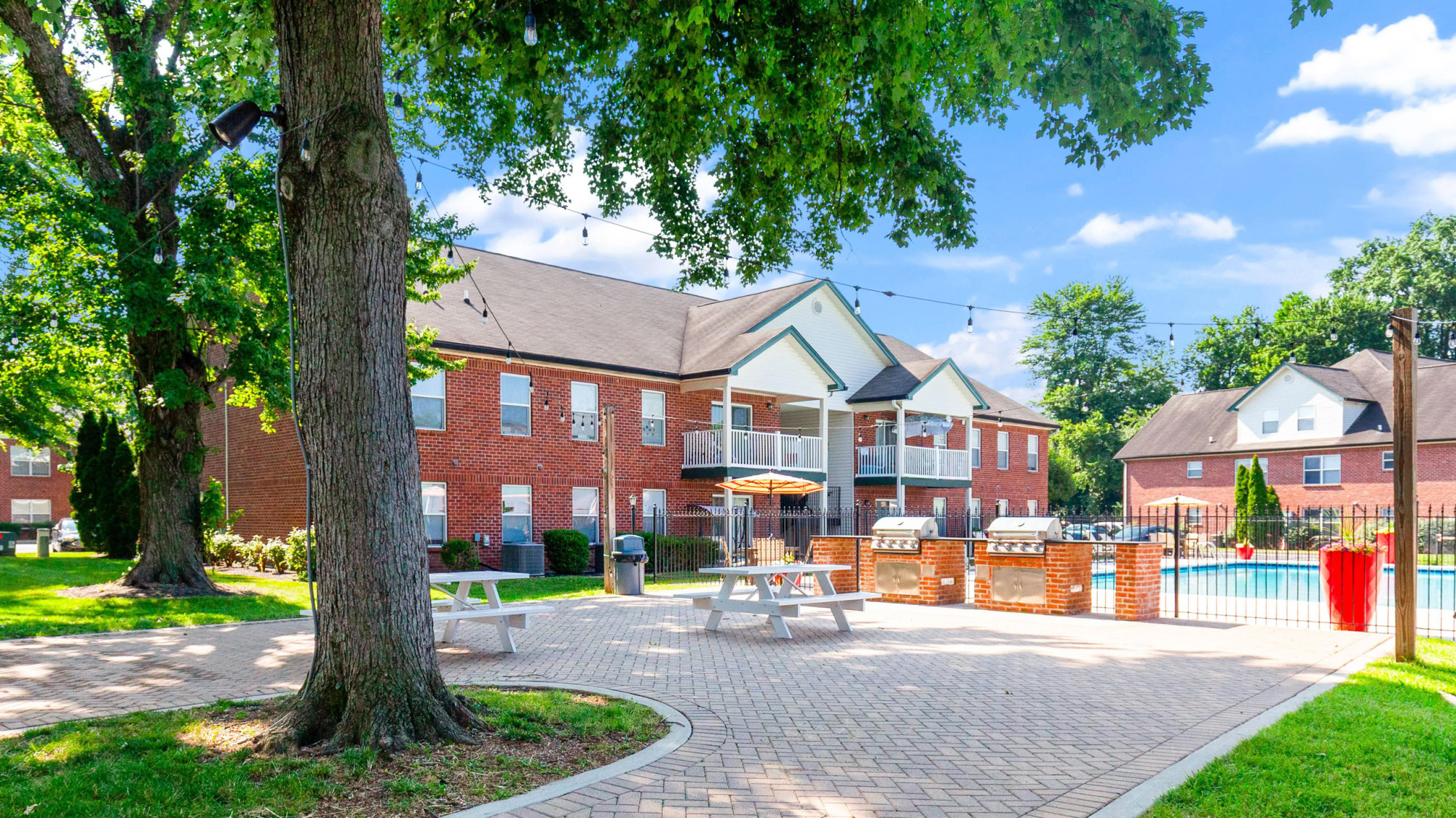 Brick apartment community exterior with white-trimmed balconies, a paved courtyard with picnic tables, mature shade trees, and a swimming pool area visible in the background.