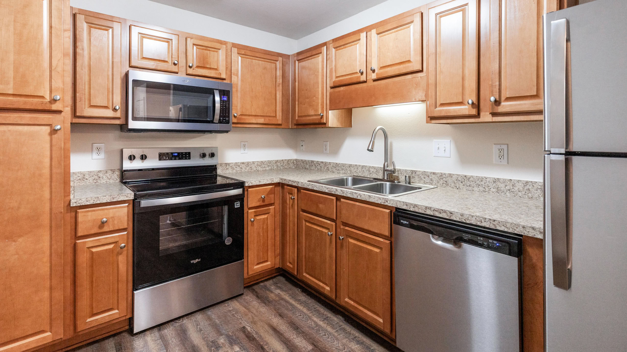 Modern apartment kitchen with warm wood cabinetry, stainless steel appliances including a microwave, electric range, dishwasher, and refrigerator, plus granite-look countertops and dark hardwood flooring.