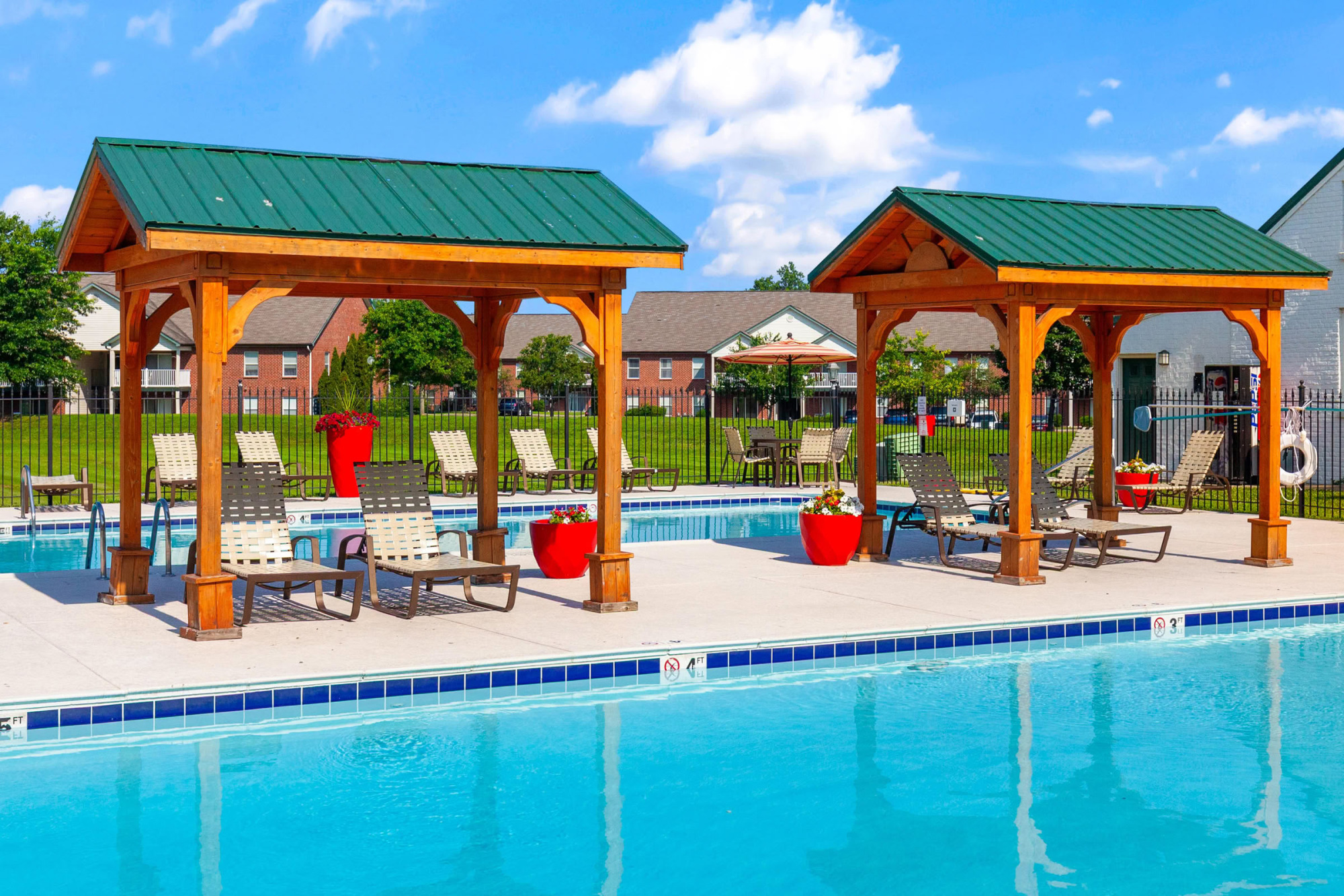 Two wooden pergolas with green metal roofs shading poolside lounge chairs and red potted flowers next to a clear blue swimming pool on a sunny day.
