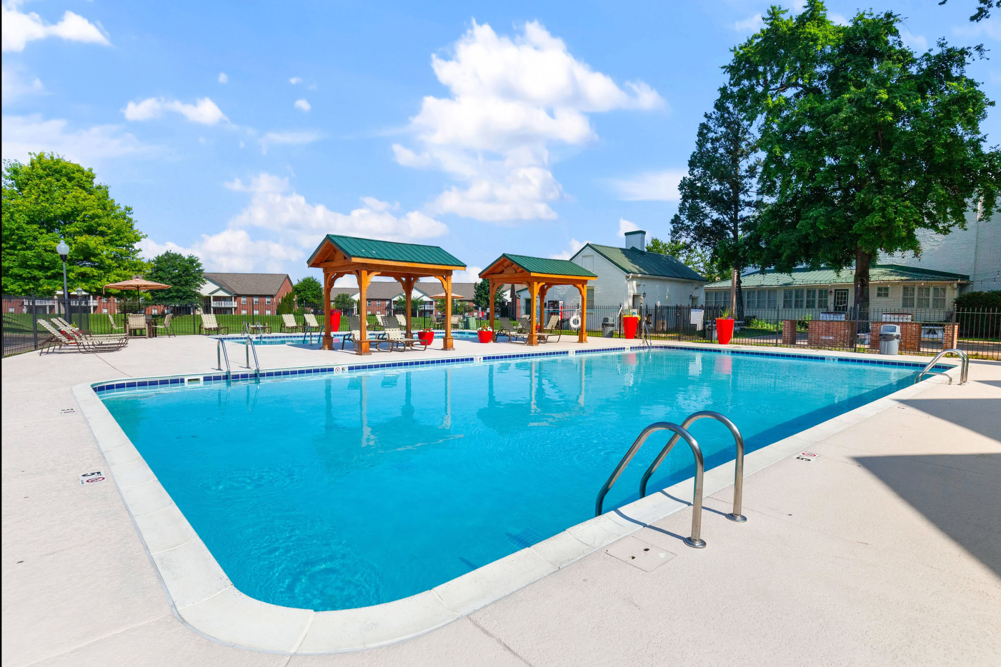 Sparkling blue resort-style swimming pool with two wooden pergolas, lounge chairs, and bright red planters set against a backdrop of trees and apartment buildings under a blue sky.