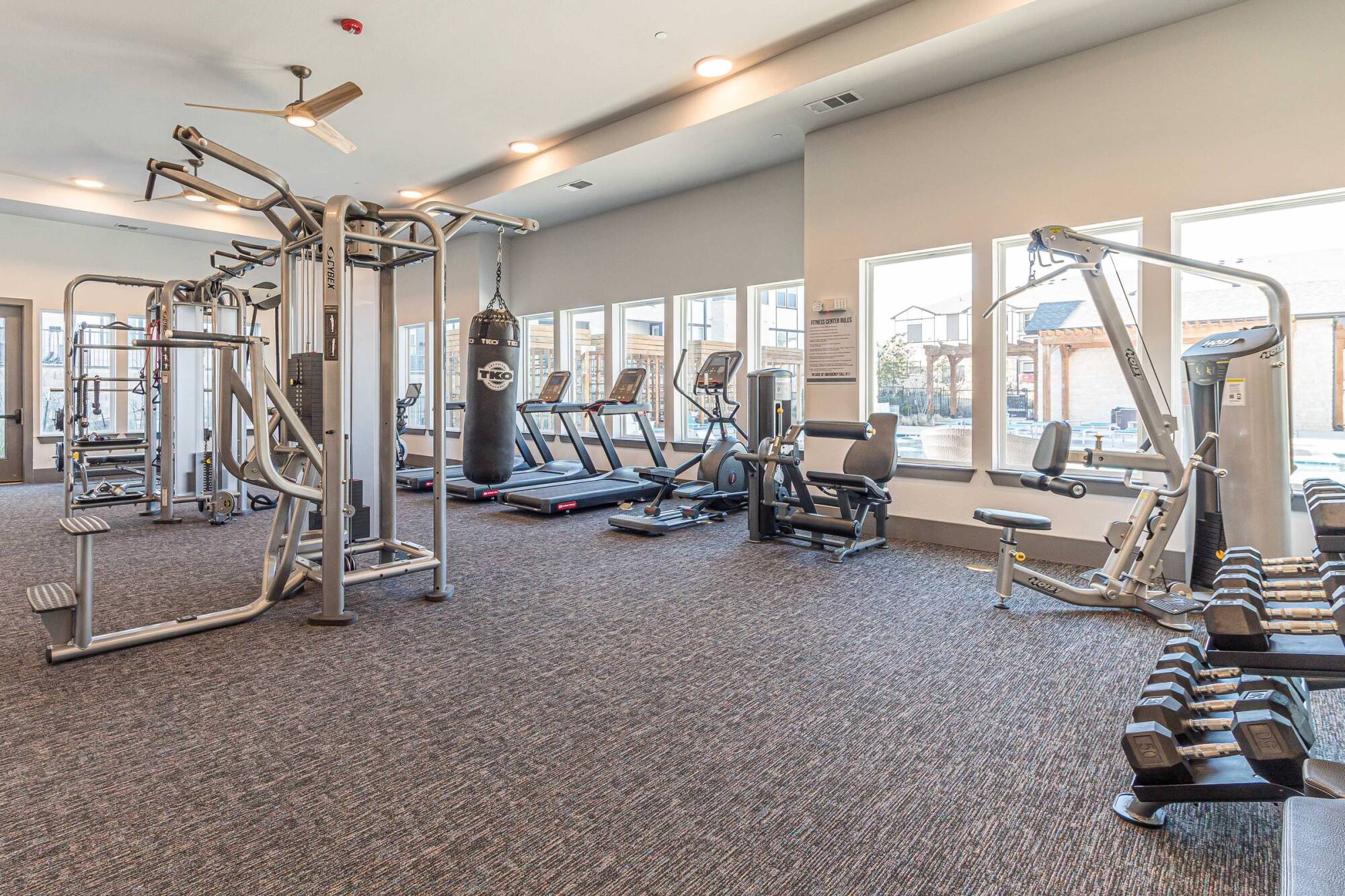 Modern gym with cardio machines, weight equipment, a punching bag, mirrors, and large windows.
