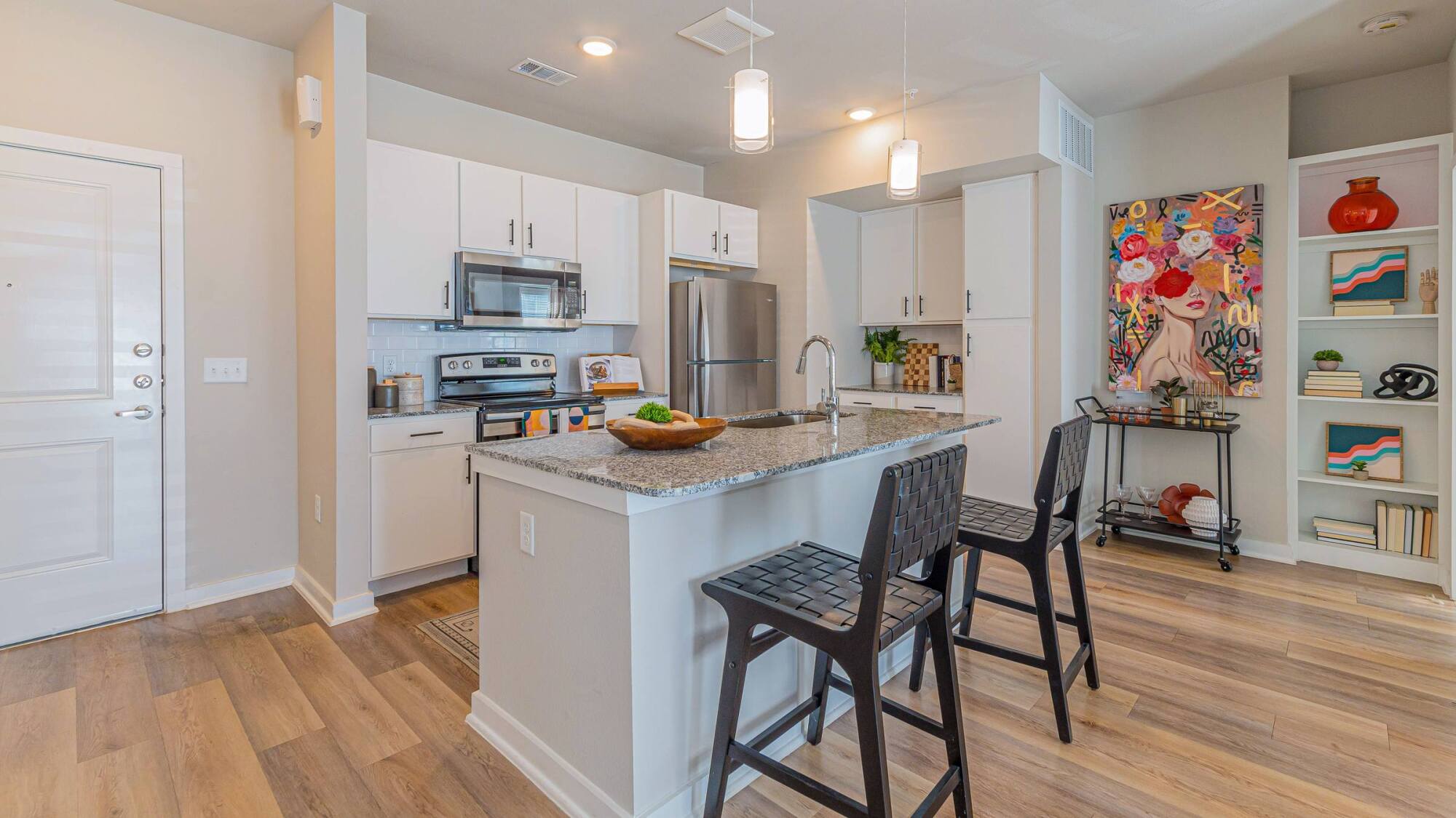 Modern kitchen with white cabinets, stainless steel appliances, island with two chairs, and colorful wall art.