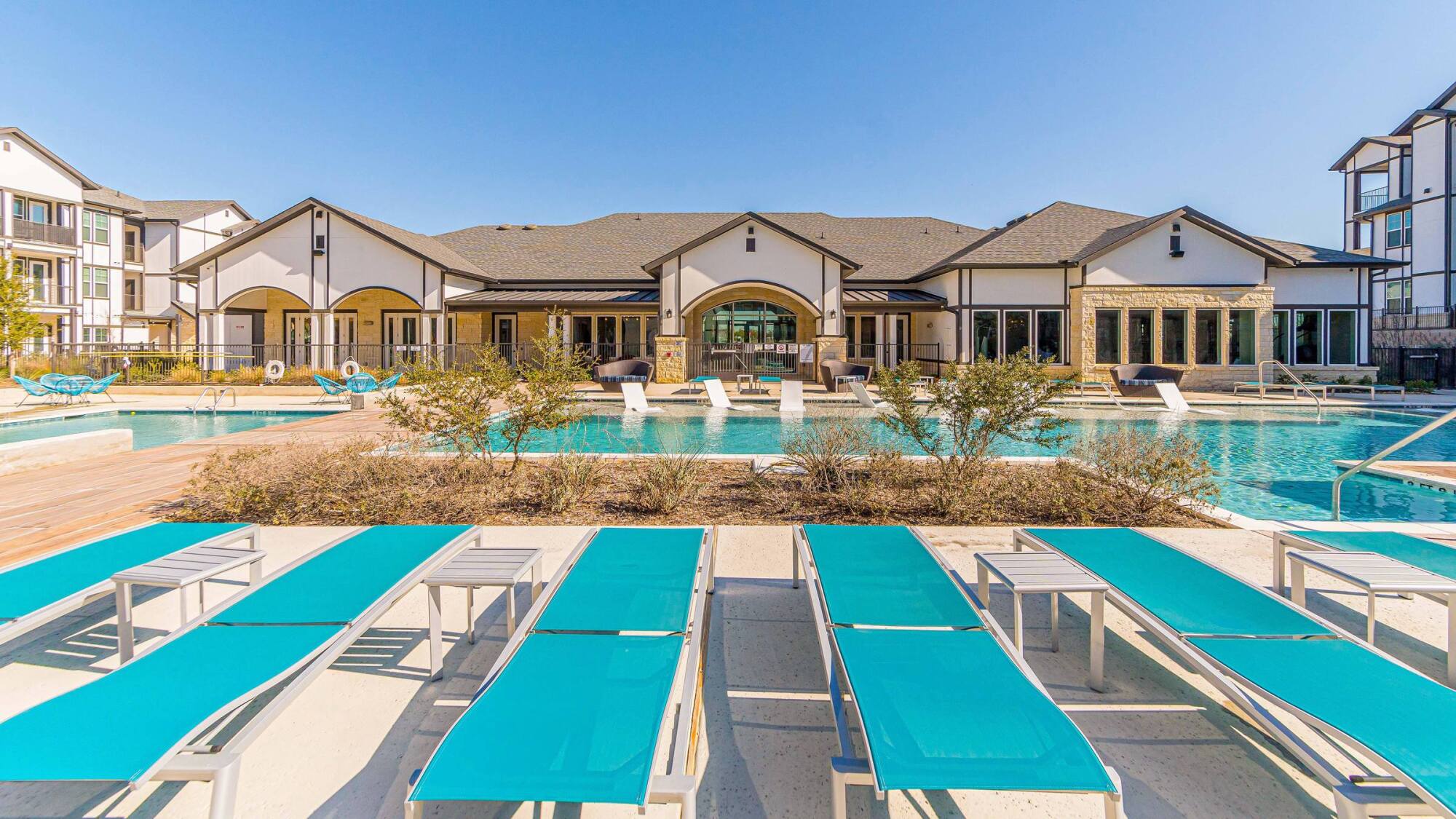 Lounge chairs by a swimming pool in front of a modern apartment complex under a clear blue sky.