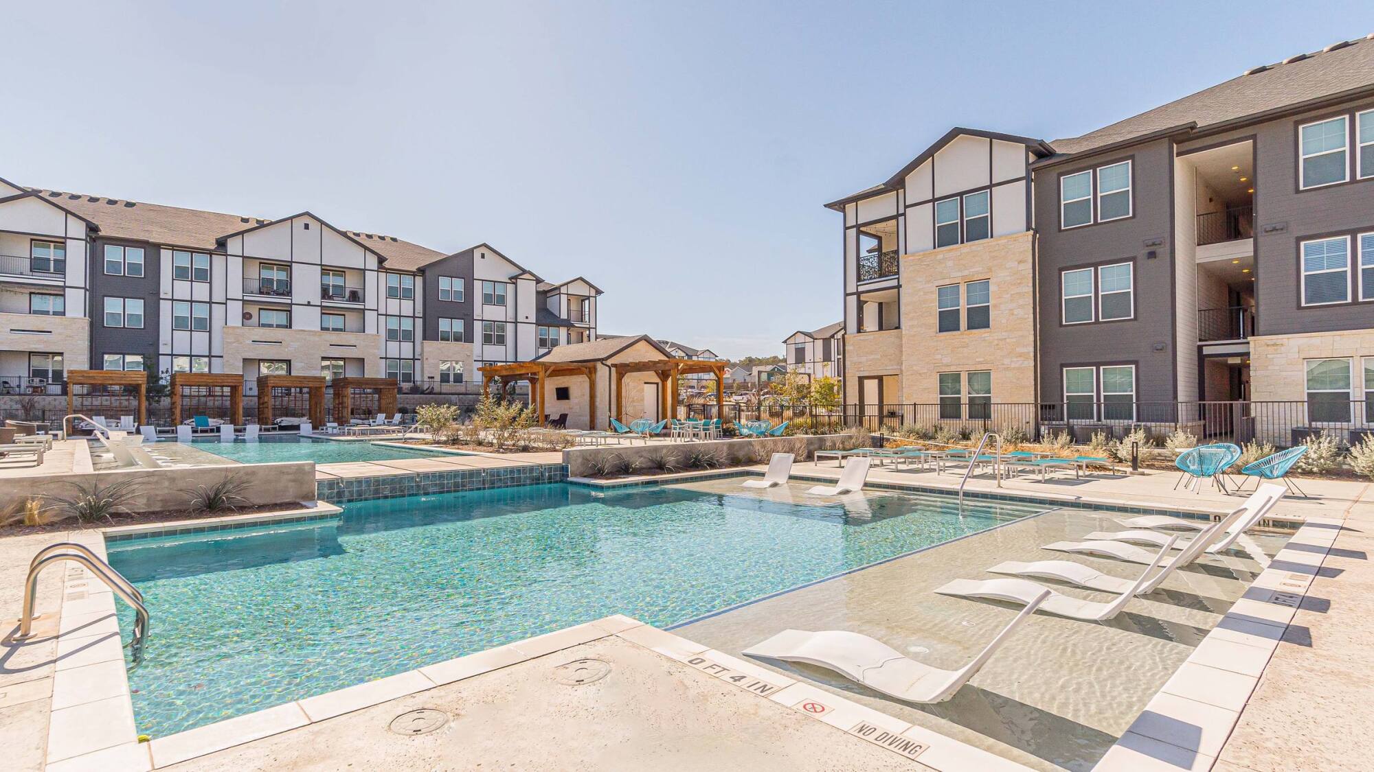 Modern apartment complex with a swimming pool, lounge chairs, and covered seating area on a sunny day.